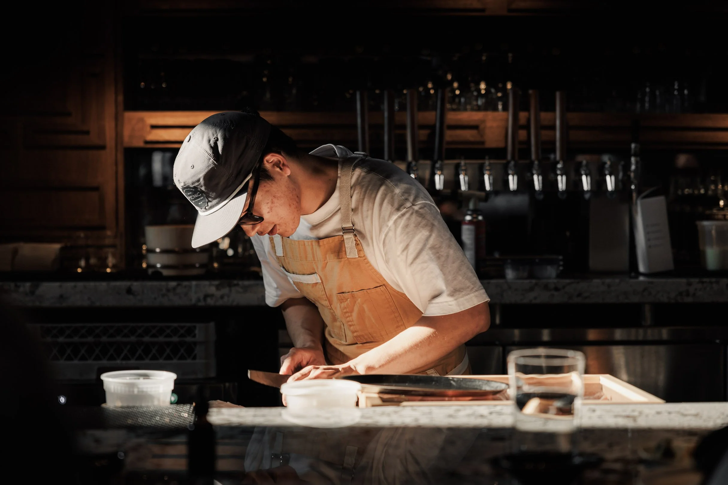 A person wearing glasses, a cap, and an apron is working in a kitchen, focusing on preparing food on a countertop with various kitchen items around.