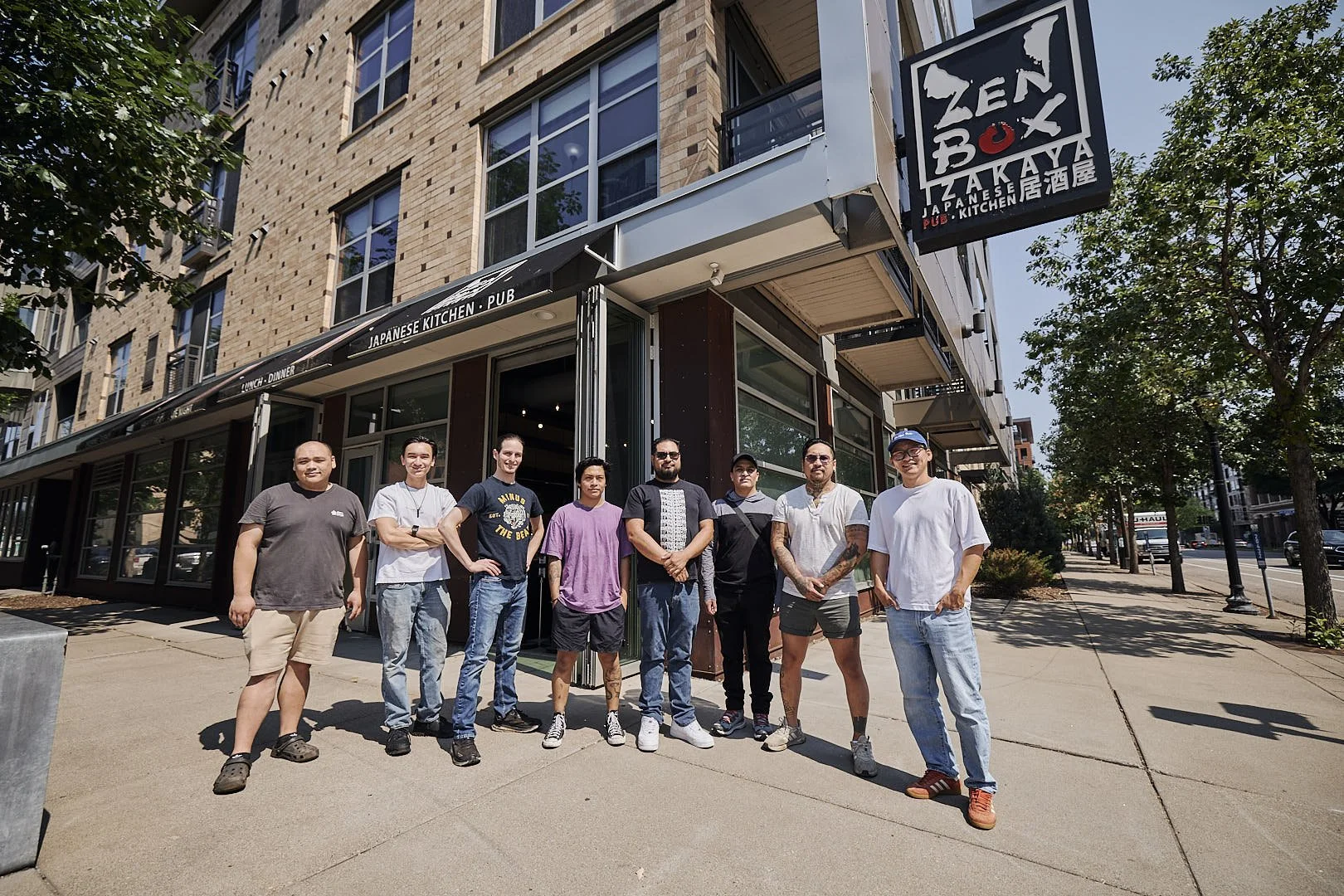 A group of nine diverse people standing outside a Japanese restaurant called Zenbox Takaya, on a city sidewalk with trees and buildings in the background.