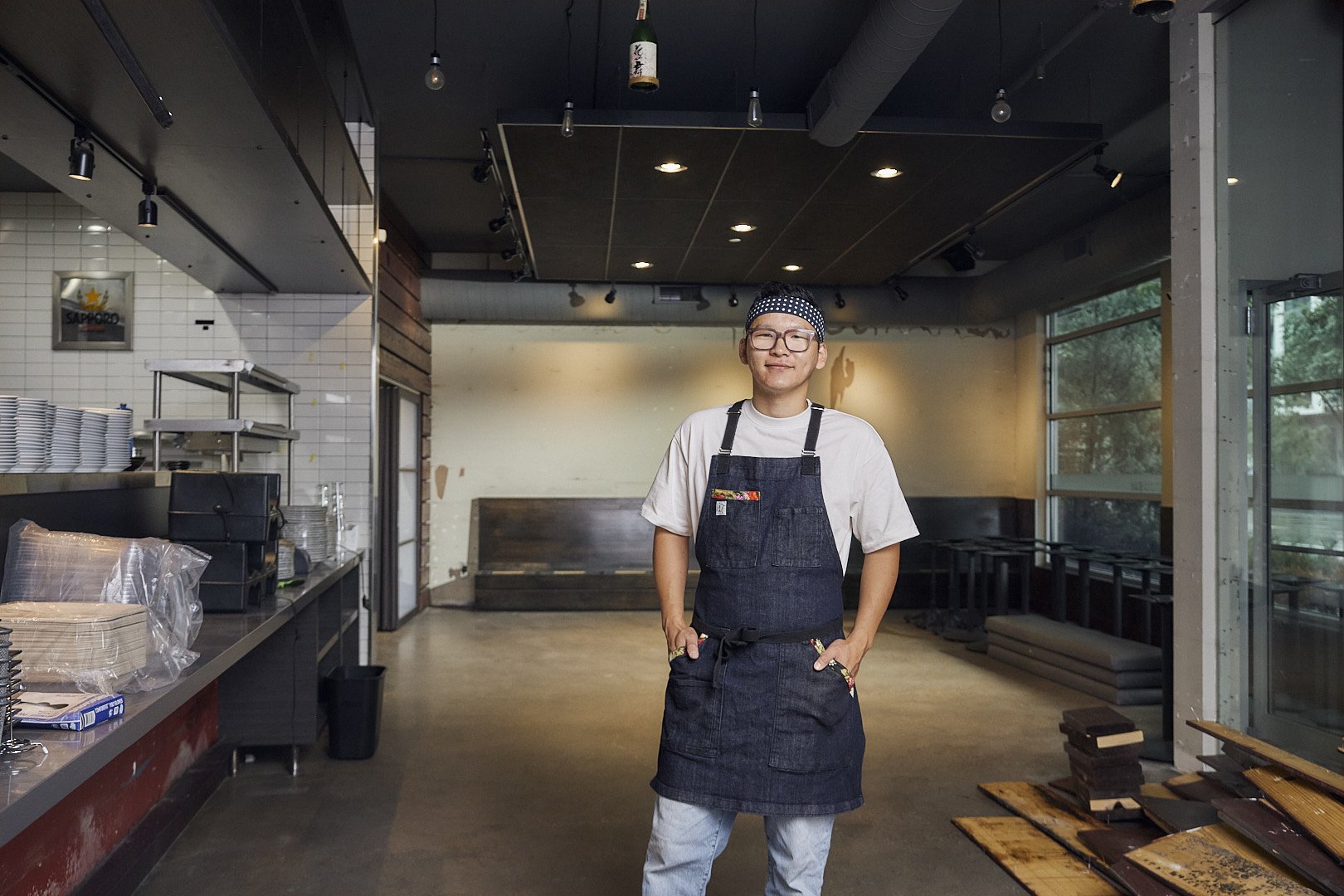 A young man wearing glasses, a polka-dotted headband, a white t-shirt, and a denim apron stands inside a modern restaurant or cafe with a confident smile, hands in pockets.