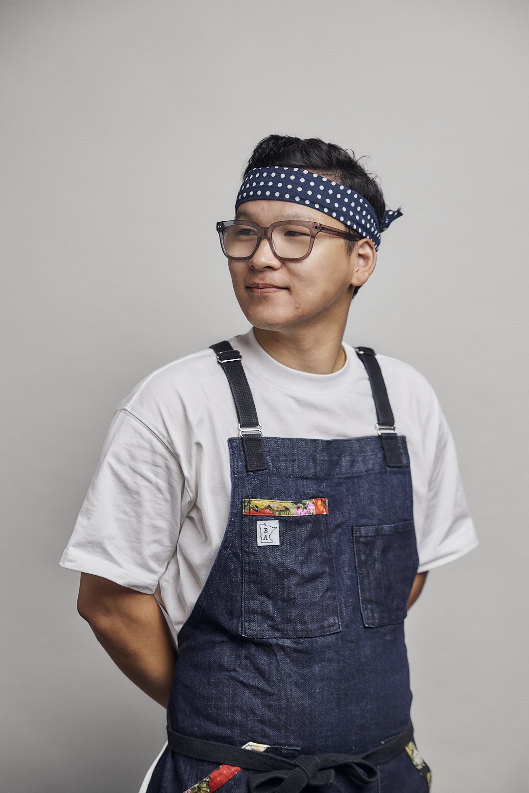A man wearing glasses, a blue polka dot headband, a white t-shirt, and a denim apron standing against a plain gray background.