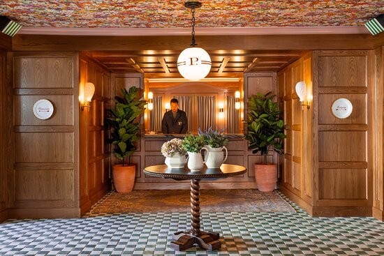 A person standing at a reception desk in a warmly lit, wood-paneled hotel lobby with potted plants and a round table with floral arrangements.