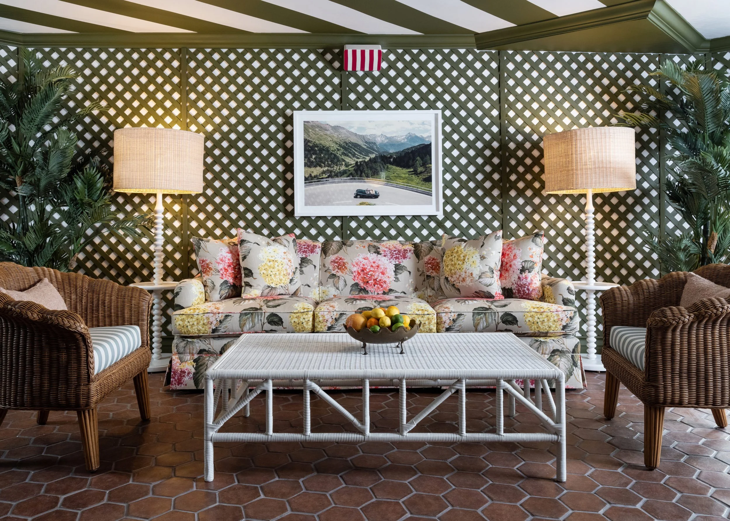 Living room with floral-patterned sofa, wicker armchairs with striped cushions, white coffee table with bowl of fruit, two tall lamps, green plants, lattice wall, framed mountain landscape photo.