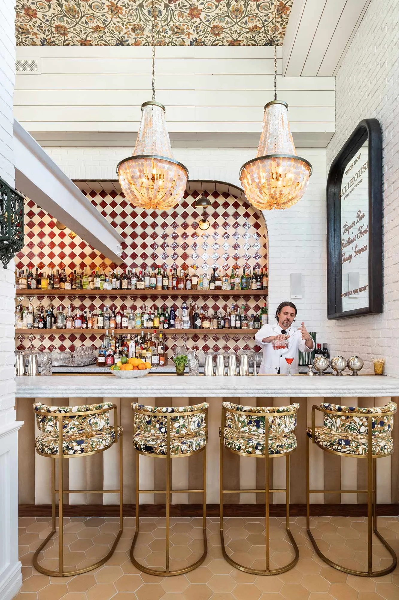 A bartender preparing a cocktail behind a marble-topped bar with four floral upholstered bar stools. The bar area features a red and white diamond-patterned tiled wall, and the bar is stocked with various bottles of alcohol. Two large hanging chandeliers with beads, a fruit bowl, and a small potted plant are on the bar counter. A large mirror with a black frame is on the right wall.