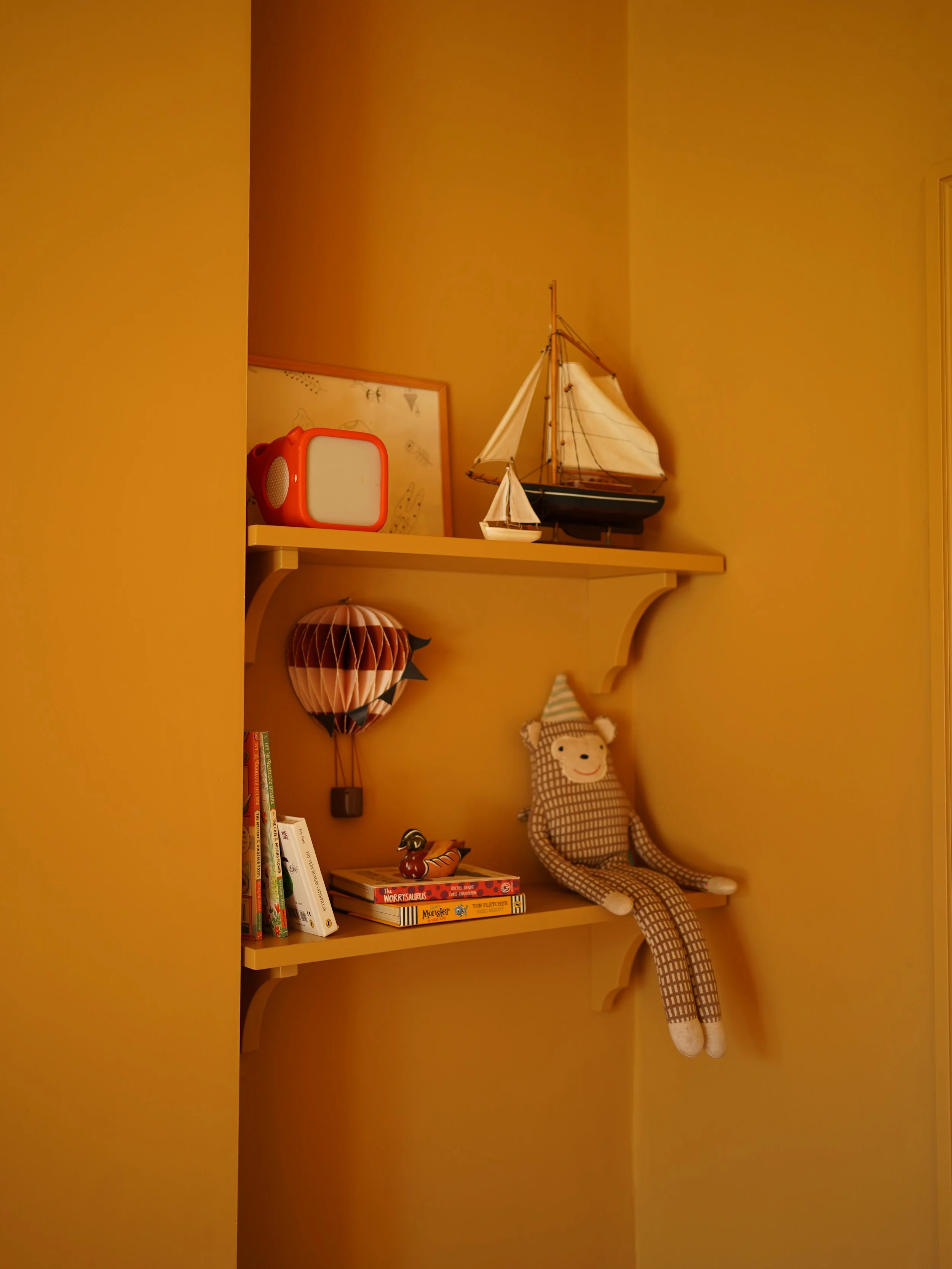A wall shelf with books, a plush doll with a lion face, a paper lantern, a model sailboat, a red portable speaker, and a framed picture.