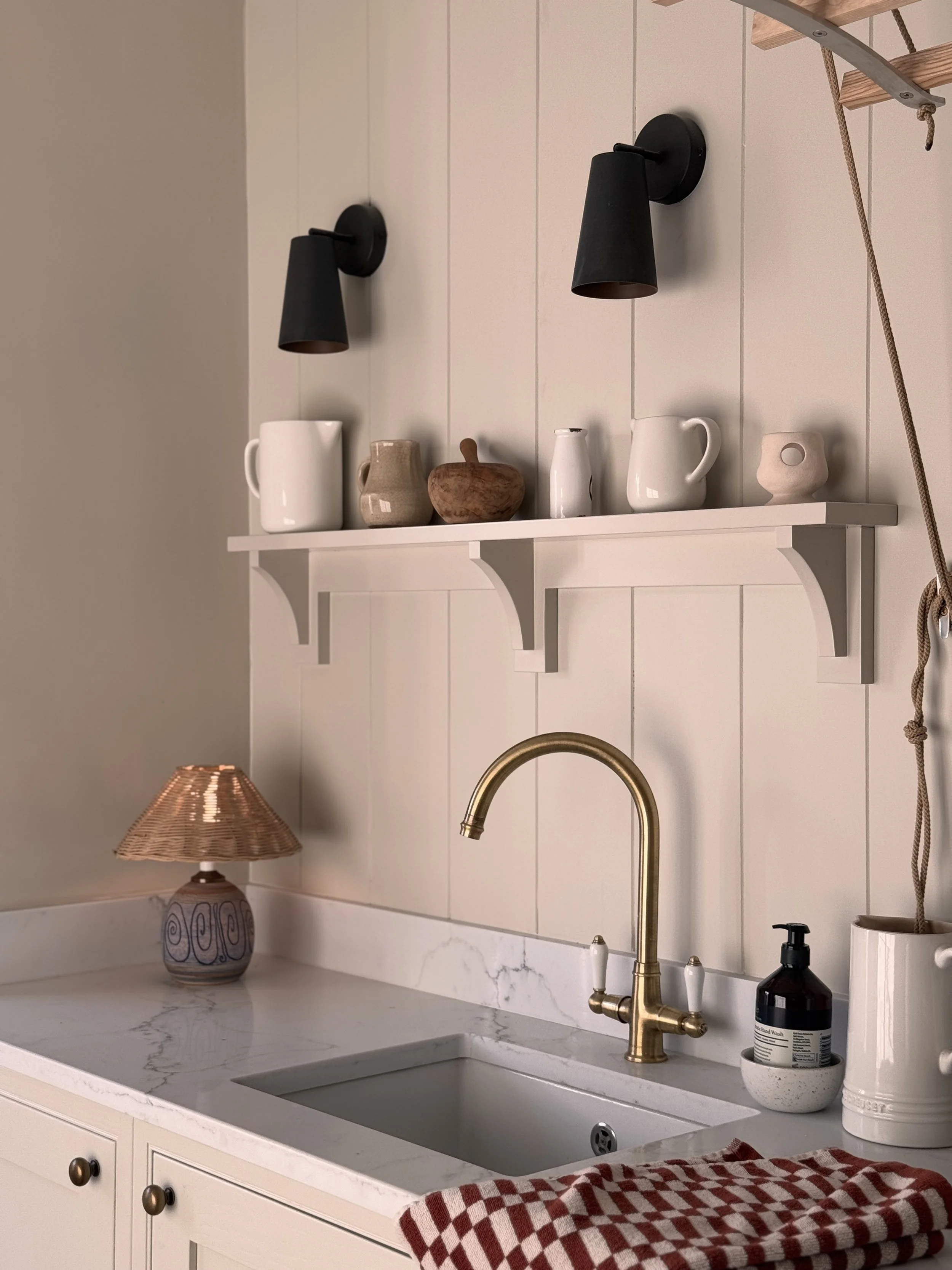 A kitchen sink with a gold faucet, white cabinetry, a marble countertop, decorative ceramic pitcher, small ceramic pot, and a soap dispenser. There are black wall-mounted lamps and a white shelf with ceramic vessels and wooden decor.