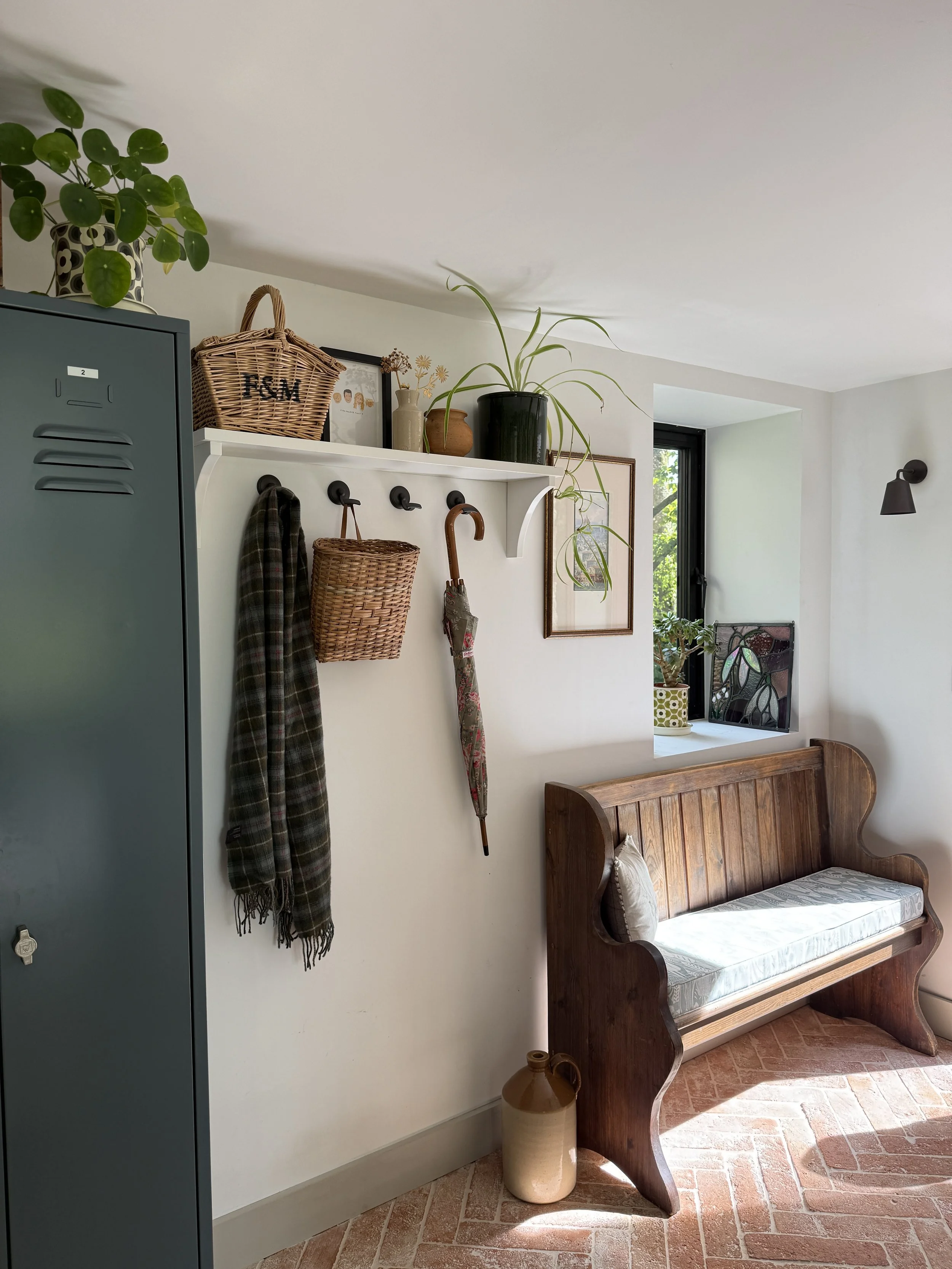 A corner of a room with a wooden bench with a cushion, a tall beige vase on the floor, a wall-mounted white shelf with plants and picture frames, two hanging baskets and clothing hooks with a plaid scarf and an umbrella, a grey locker, and a window with a view of greenery outside.