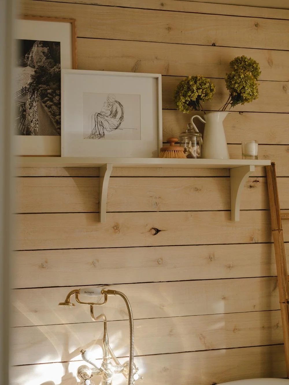 Shelf with framed art, potted plant, jars, candle, and wooden stick against a wooden wall, with vintage faucet and sink below.