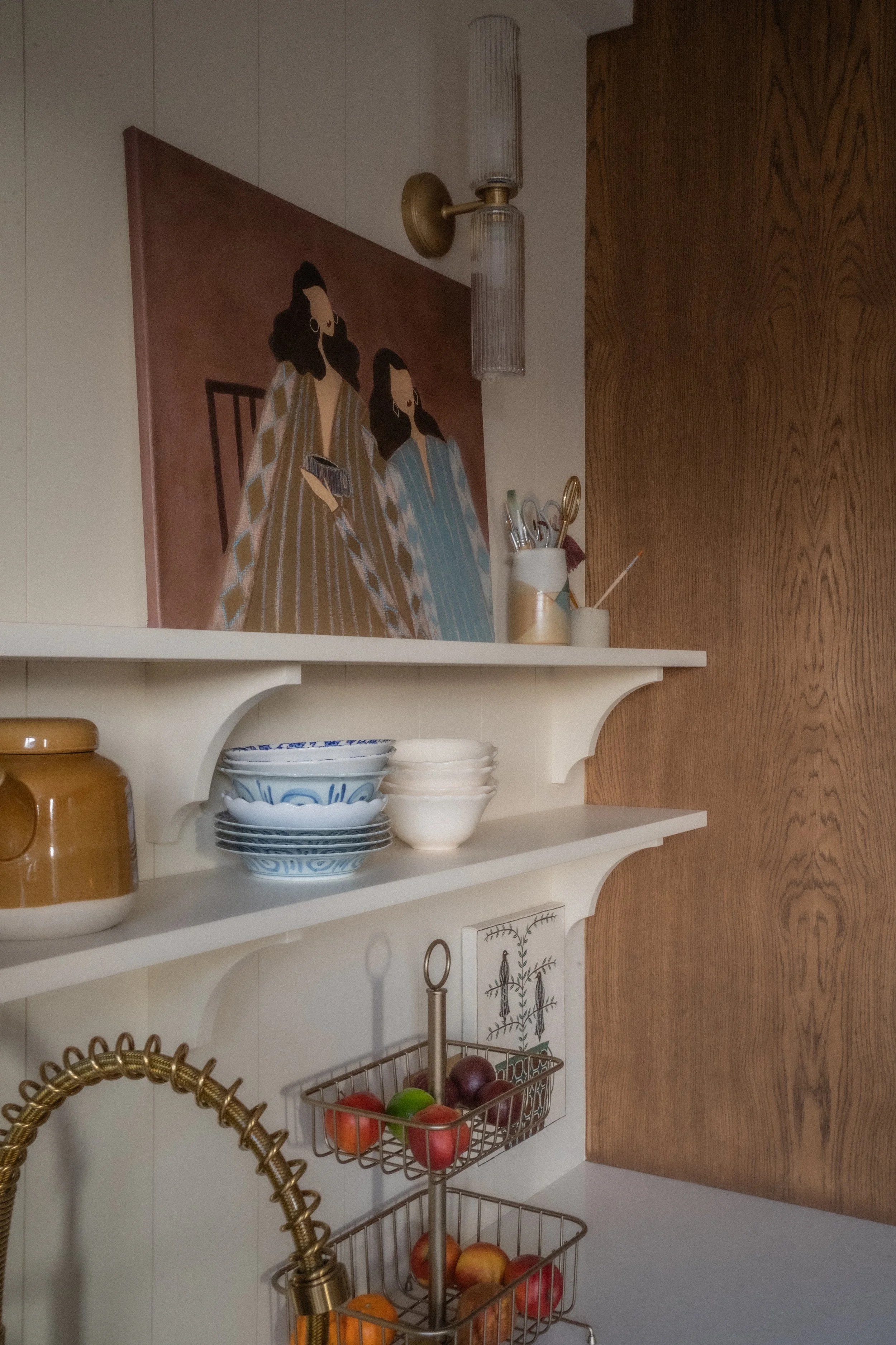 Kitchen shelf with ceramic bowls, a pitcher, and a jar, above a fruit basket and gold sink faucet, with a wall-mounted artwork of women in traditional clothing.