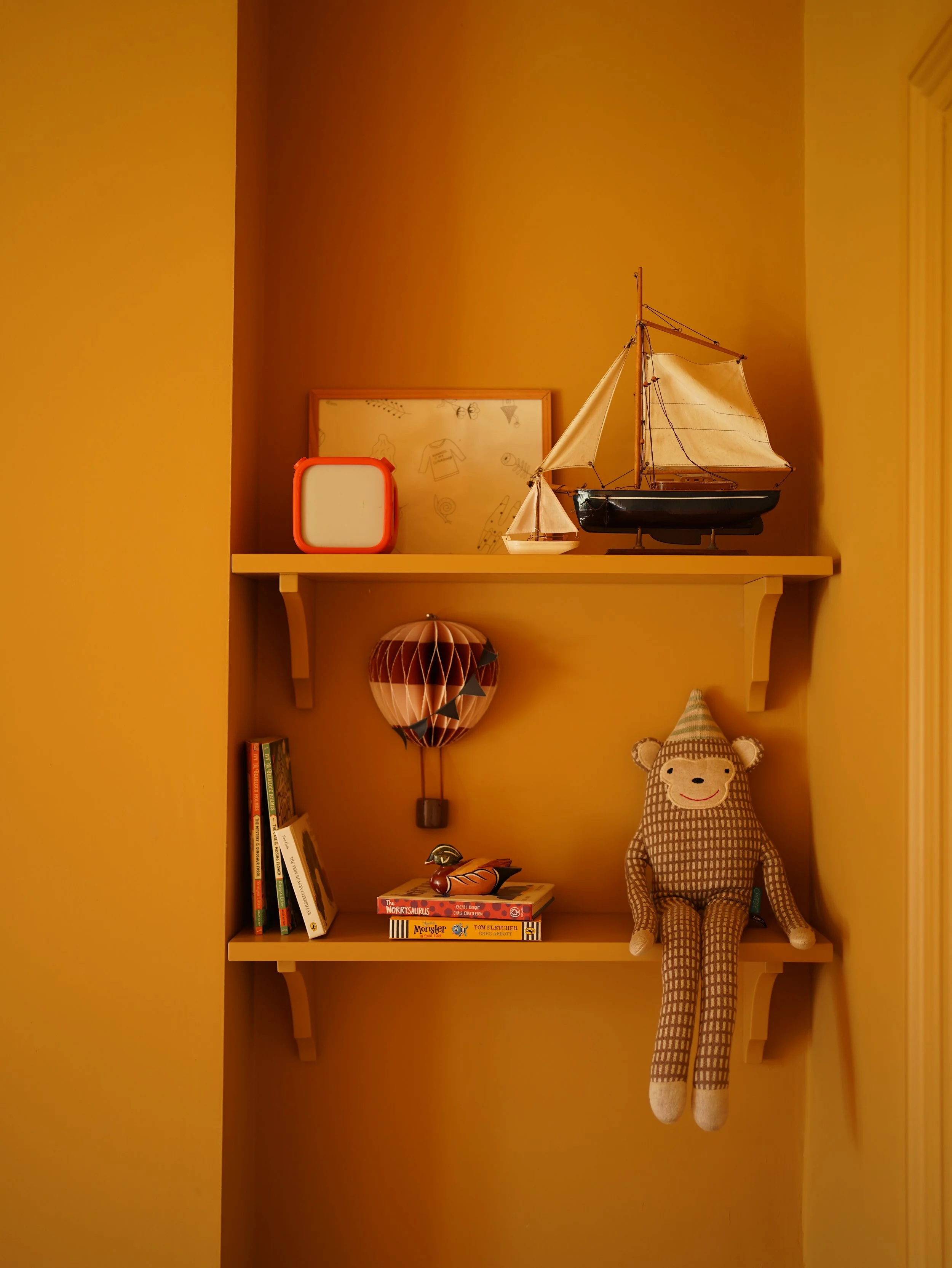 A yellow wall with a white shelf holding a model sailboat, a book-themed framed picture, a small square light, and a pile of children's books, with a plush gingerbread man and a paper lantern below.