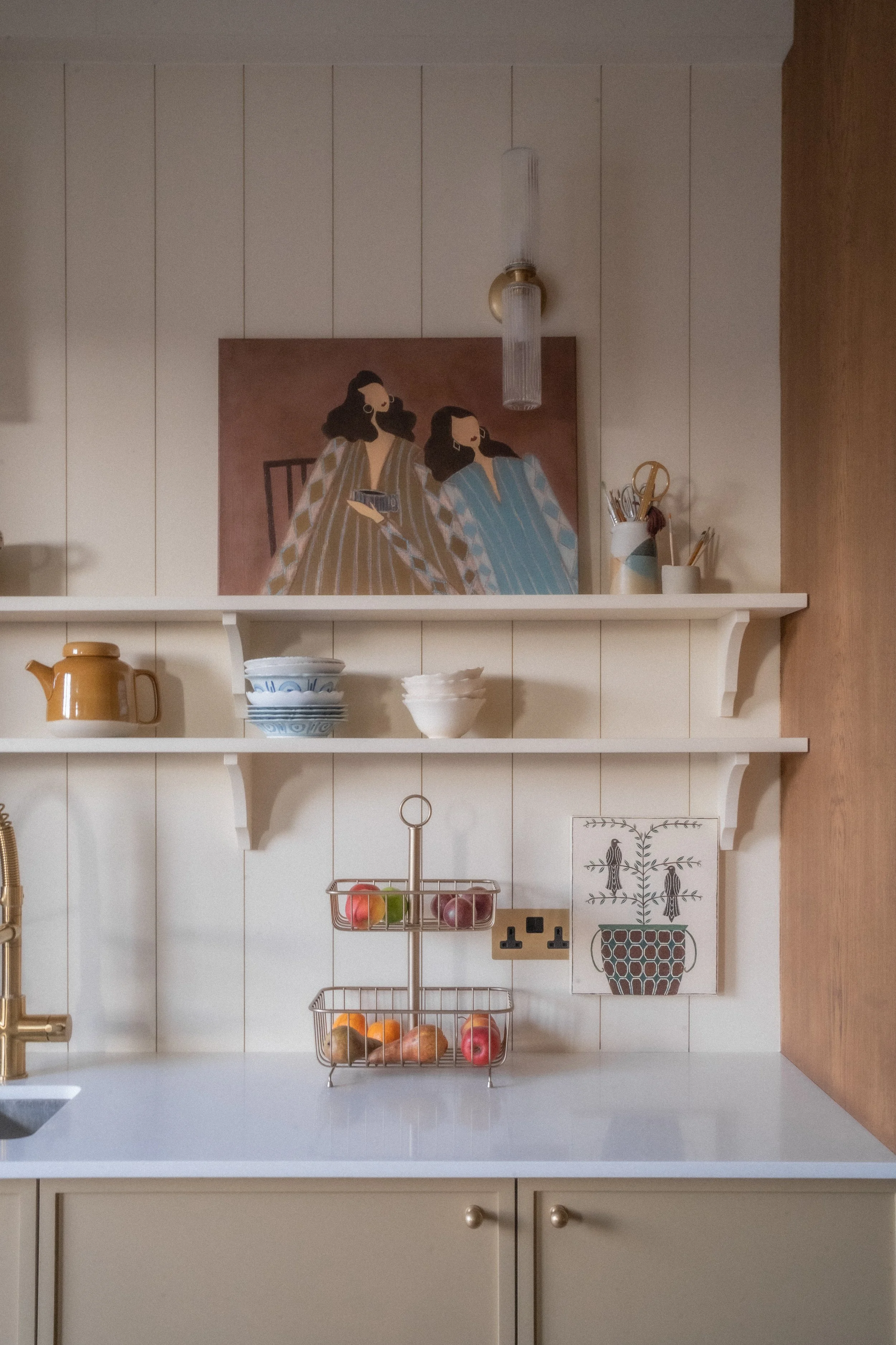 Kitchen shelf with containers and bowls, wall decor with bird illustration, fruit basket with apples and oranges, light beige cabinet, and a wall-mounted light fixture.