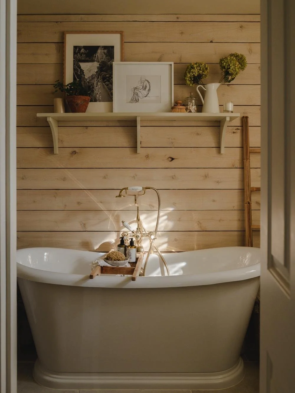 A bathroom with a vintage bathtub in front of a wood-paneled wall, a shelf with framed art, greenery, and decorative items, and a ladder leaning against the wall.