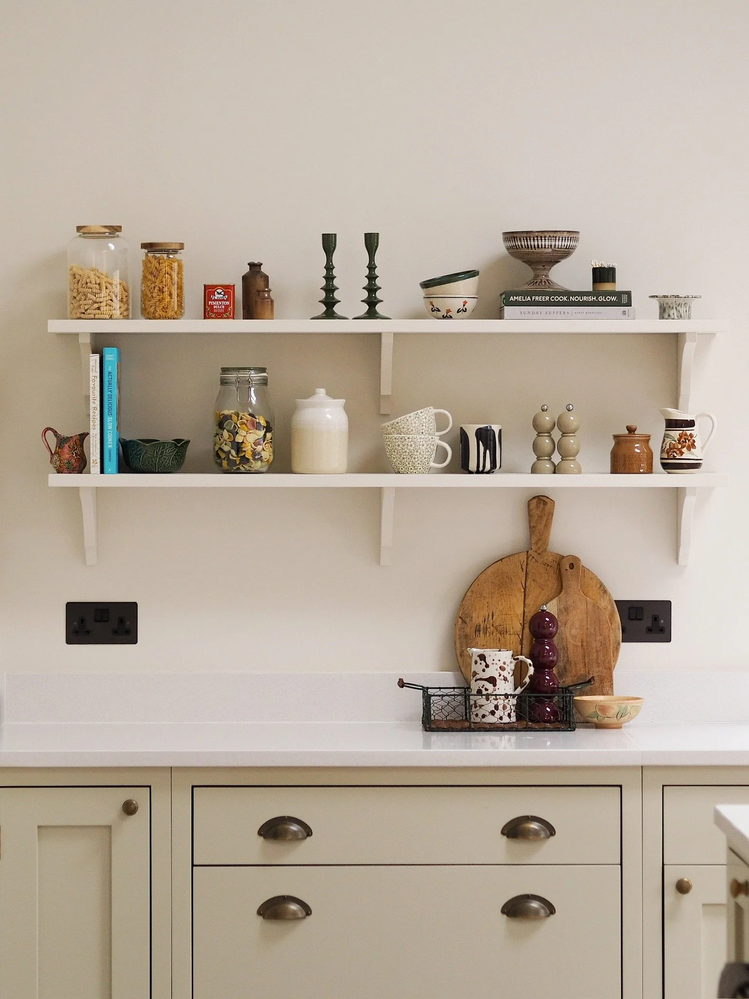 White kitchen cabinet with three drawers and two cabinets below, black electrical outlets on the wall behind, decorated with various kitchenware and decorations on two white wall-mounted shelves.