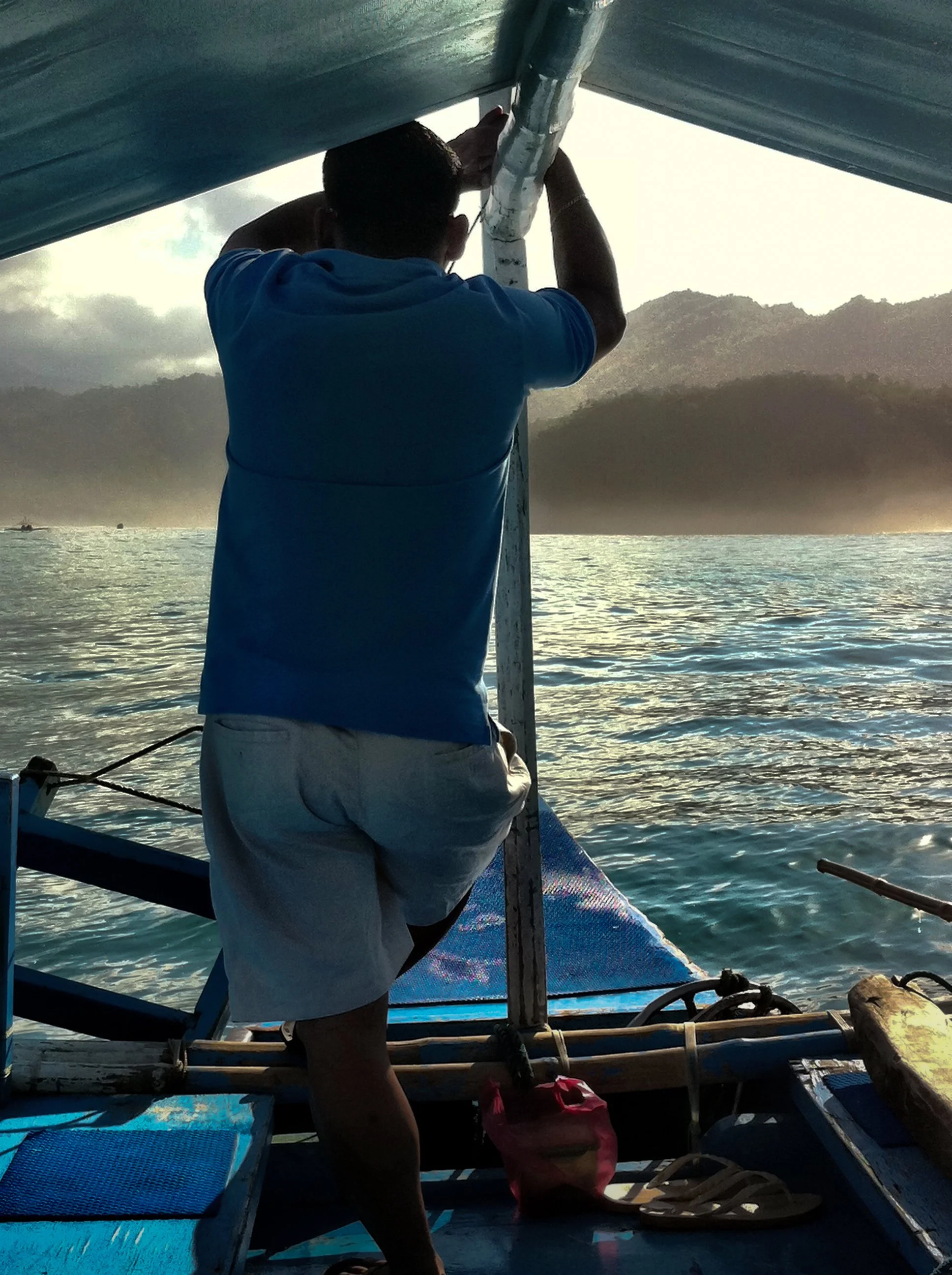 A man standing on a boat, holding onto a pole, with a lake and mountain landscape in the background at sunset.