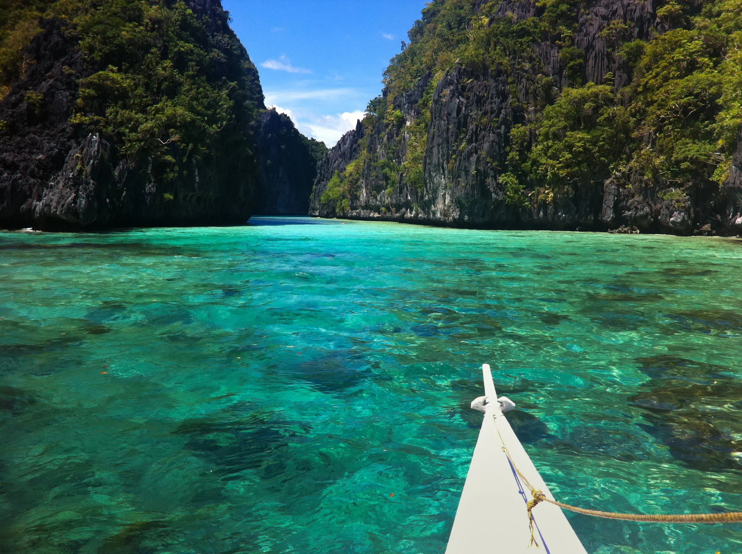 A boat's bow pointing towards clear turquoise water between lush, green, rocky cliffs with a blue sky overhead.