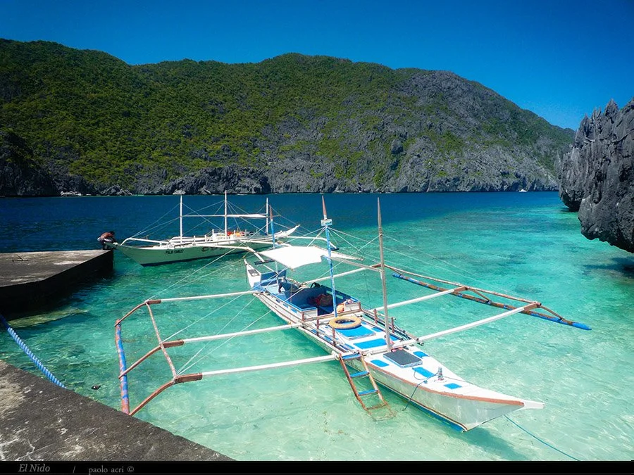 A traditional outrigger boat anchored in clear turquoise water near a rocky shoreline, with green forested hills in the background under a blue sky.