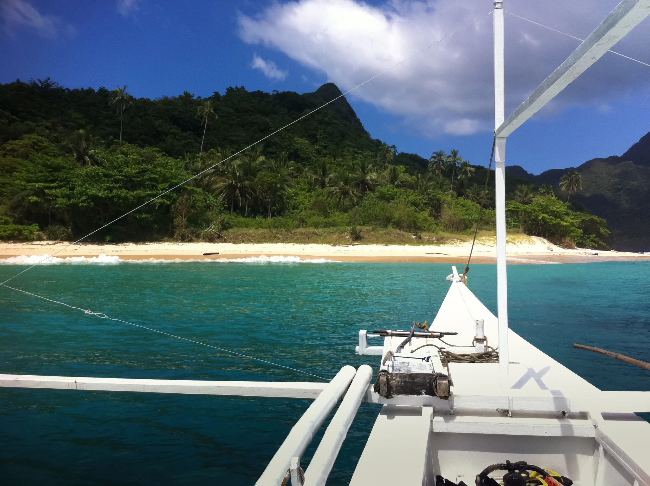 View from a boat on turquoise water approaching a tropical island with sandy beach and lush green trees, with mountains in the background under partly cloudy sky.