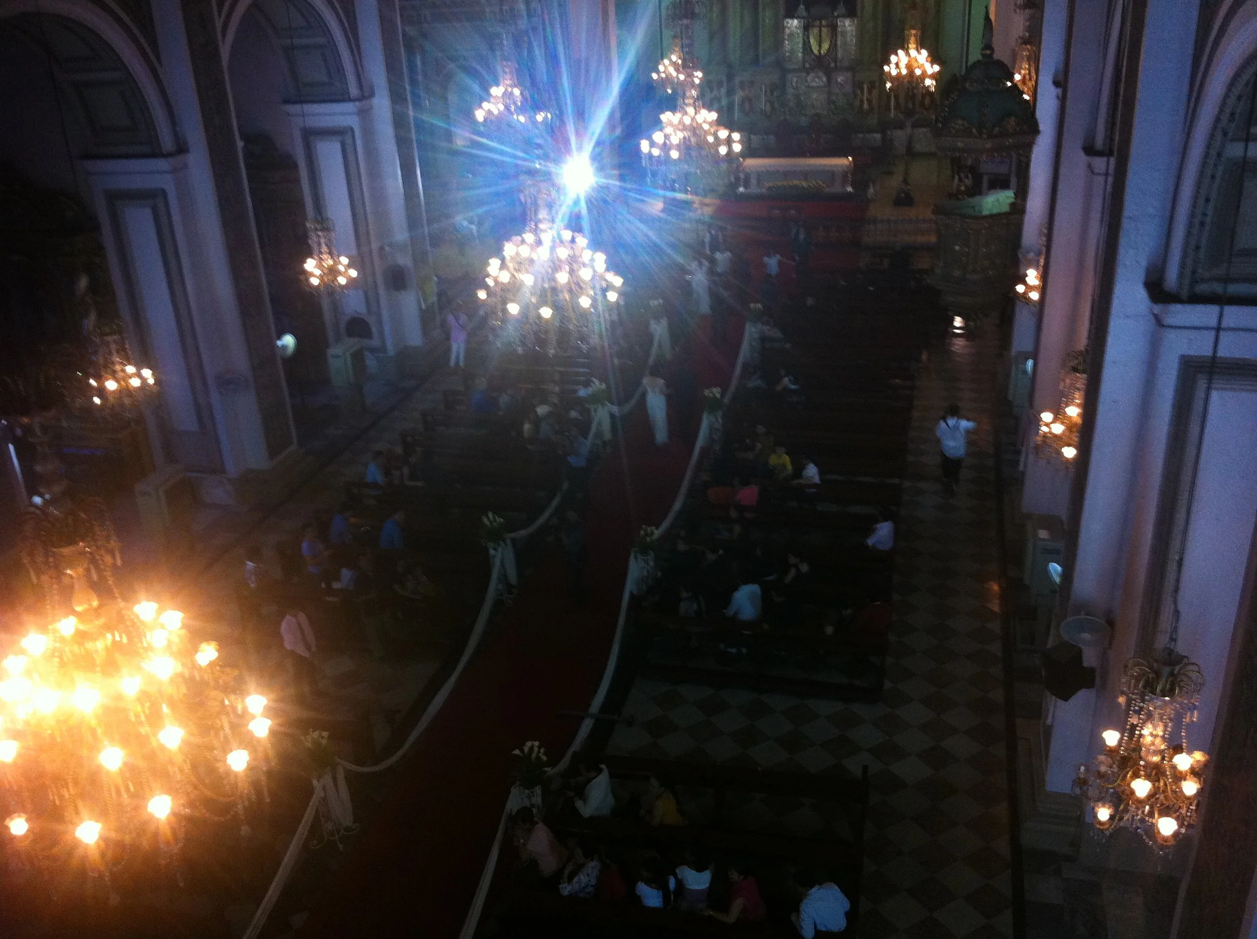 The interior of a church or cathedral with ornate chandeliers, long red carpet aisle, pews filled with seated congregants, and a bright spotlight at the front.