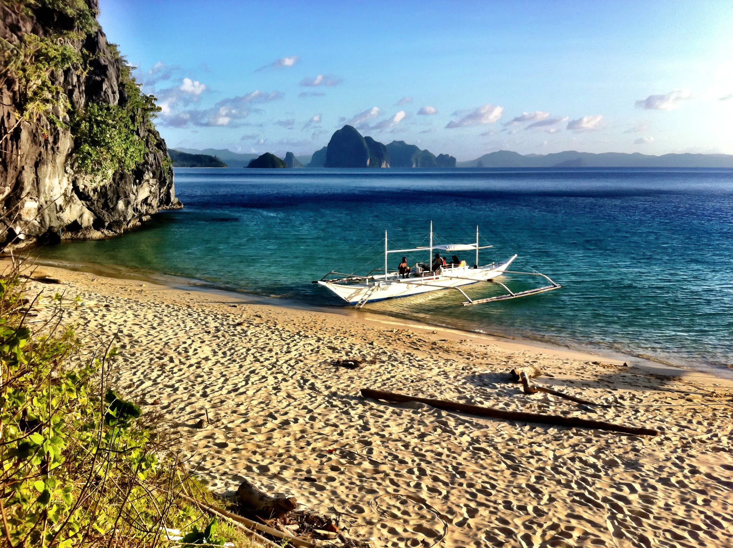 Beach with golden sand, a sailboat near the water's edge, rocky cliff on the left, distant islands, and clear blue sky with some clouds.