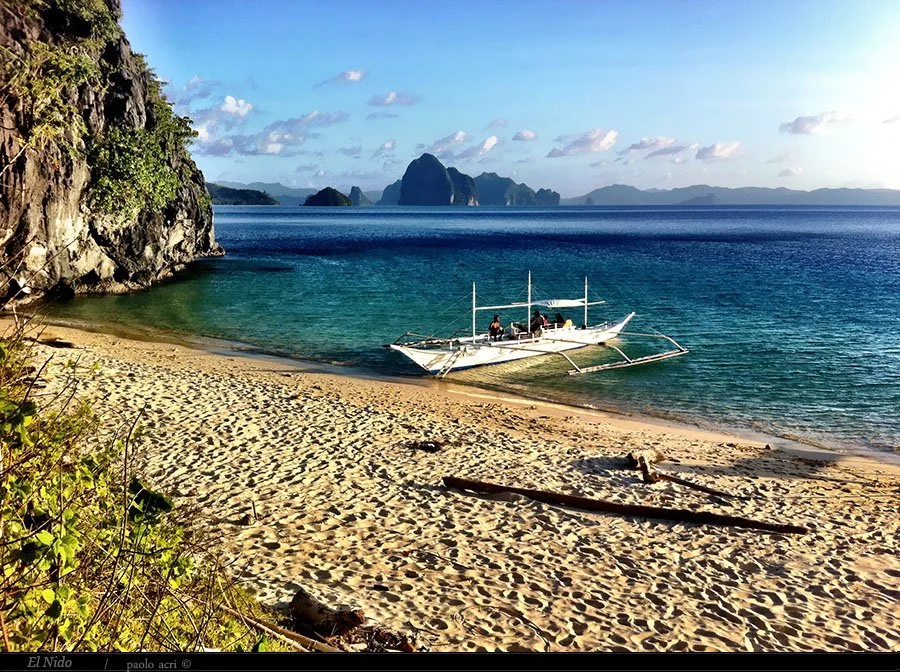 A peaceful beach with golden sand, a white boat with outriggers floating near the shore, and lush green cliffs. In the background, clear blue water extends to distant islands and mountains under a partly cloudy sky.