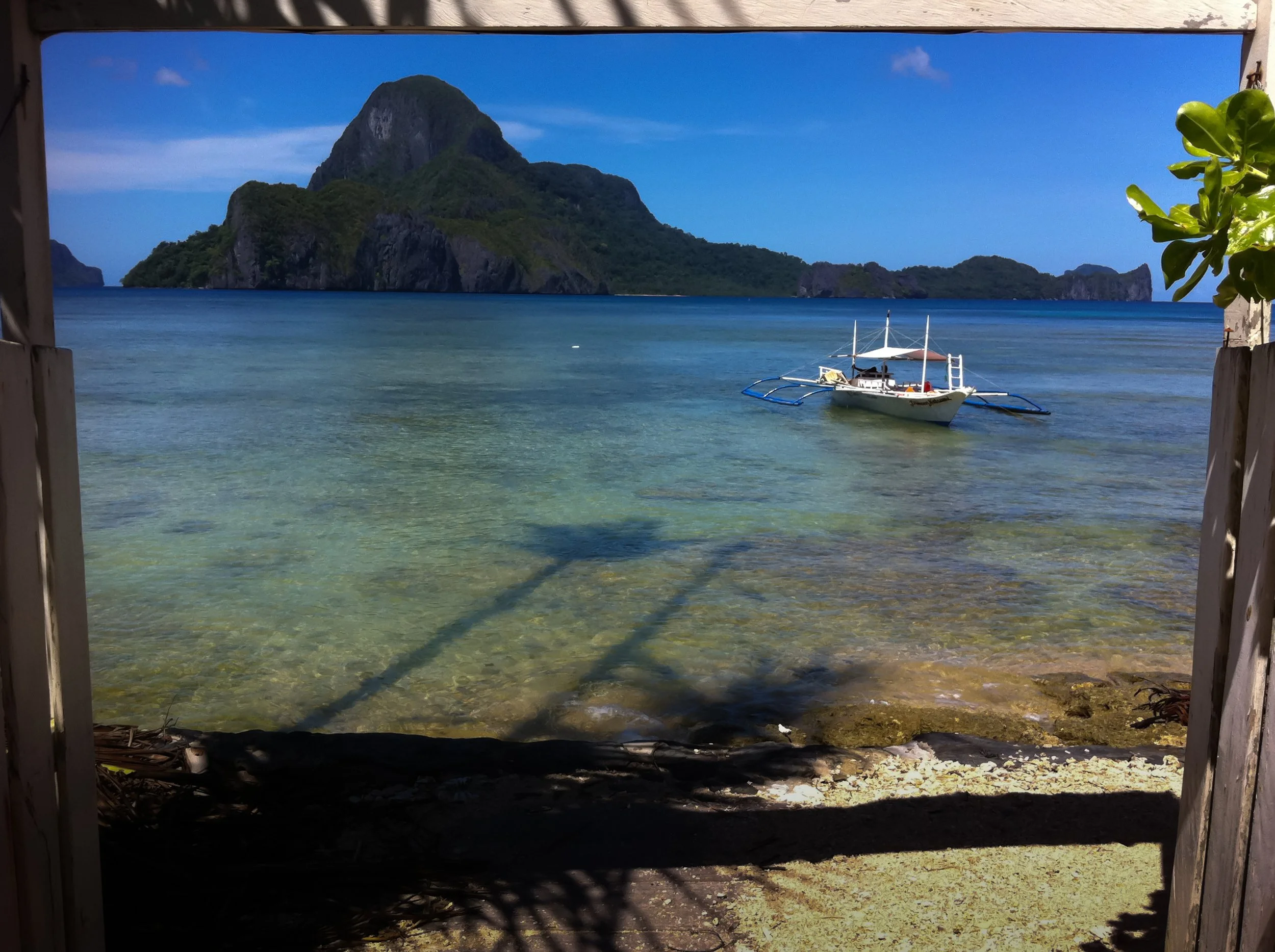 A tropical beach scene with clear blue water, a small boat floating near the shore, and lush green islands with cliffs in the background, all viewed through a wooden structure.