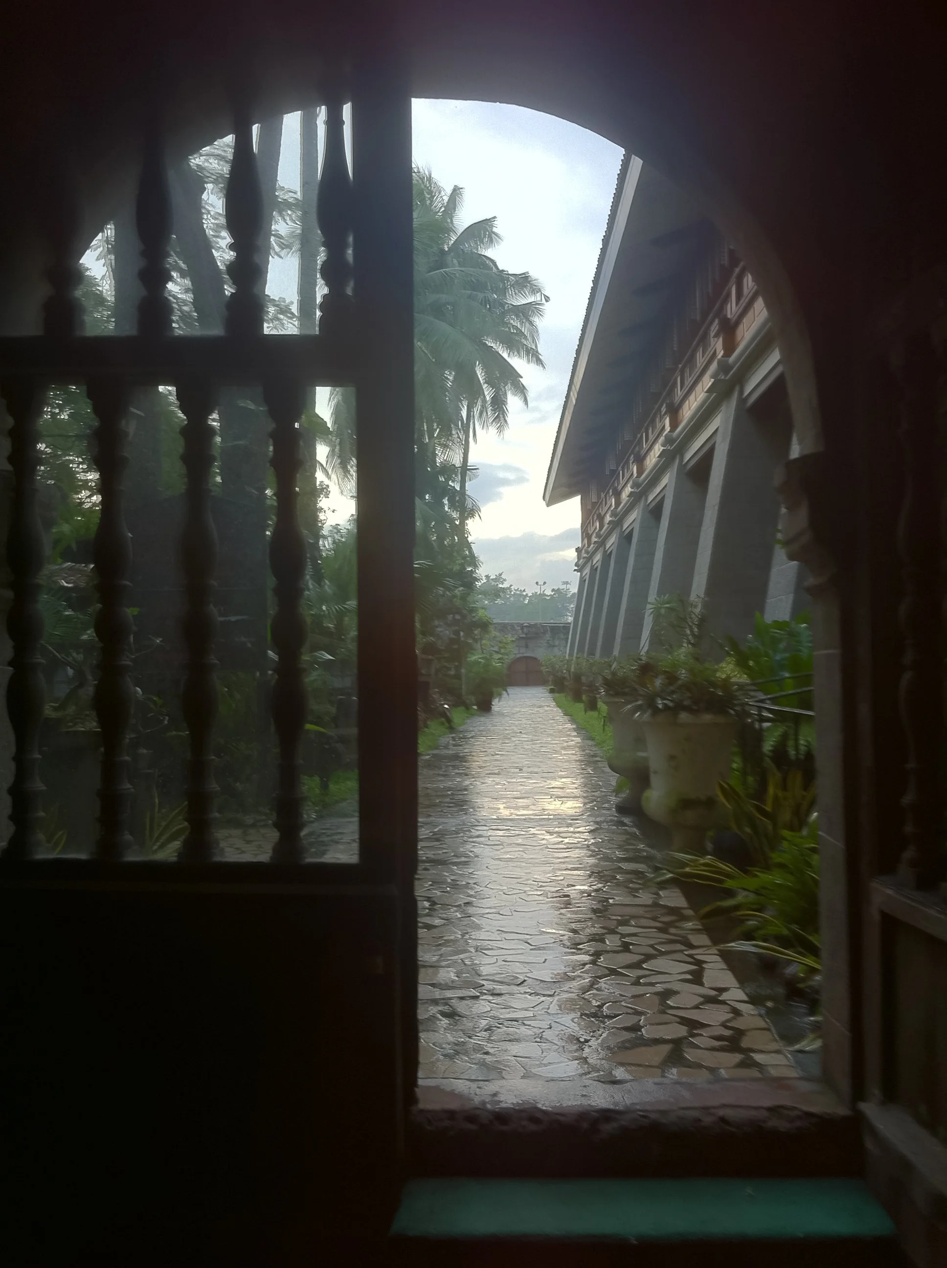 View through an arched window showing a wet stone pathway, large potted plants, palm trees, and a building with large supports, under an overcast sky.