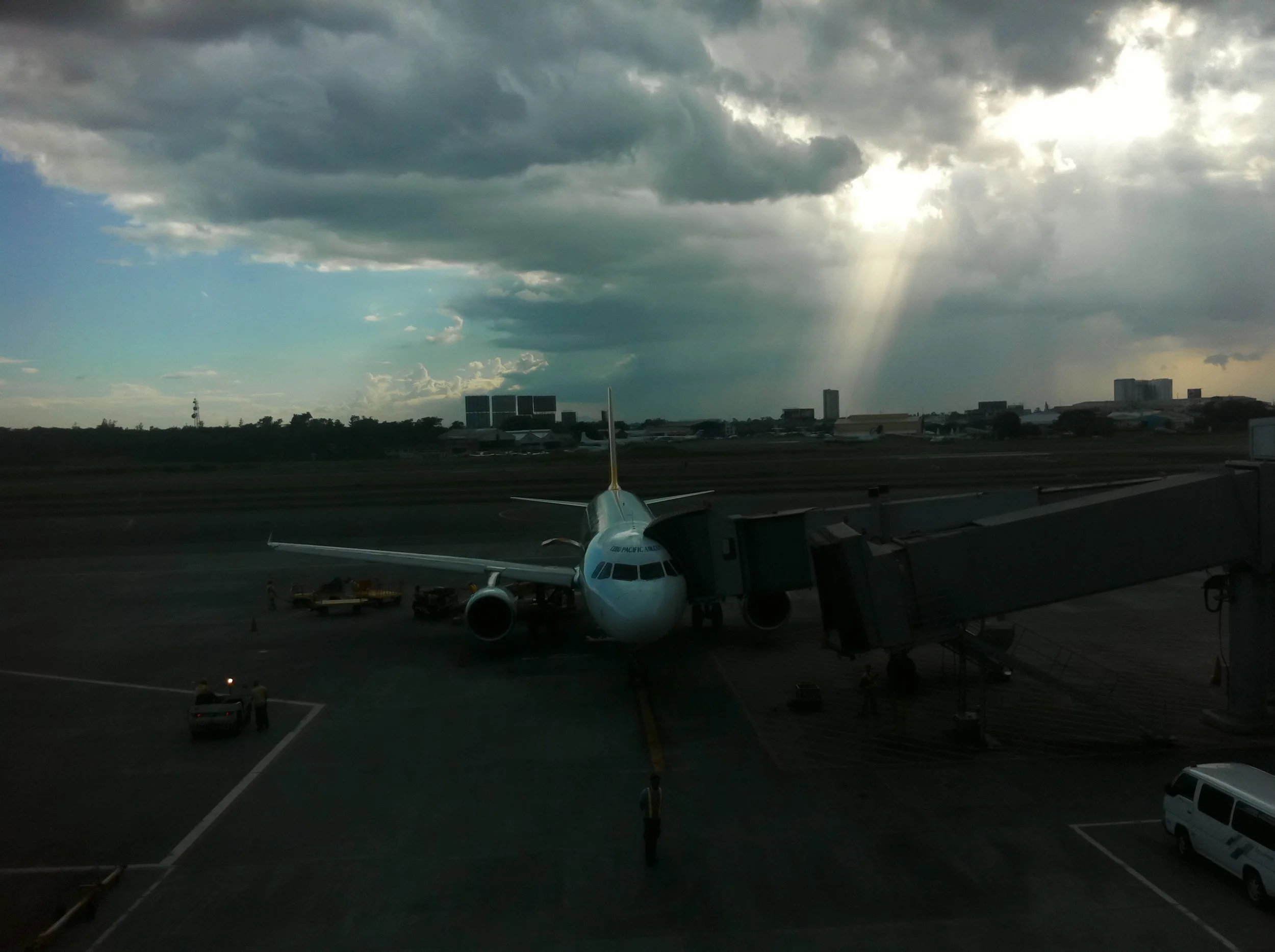 An airplane parked at an airport gate under a cloudy sky with sunlight piercing through clouds.