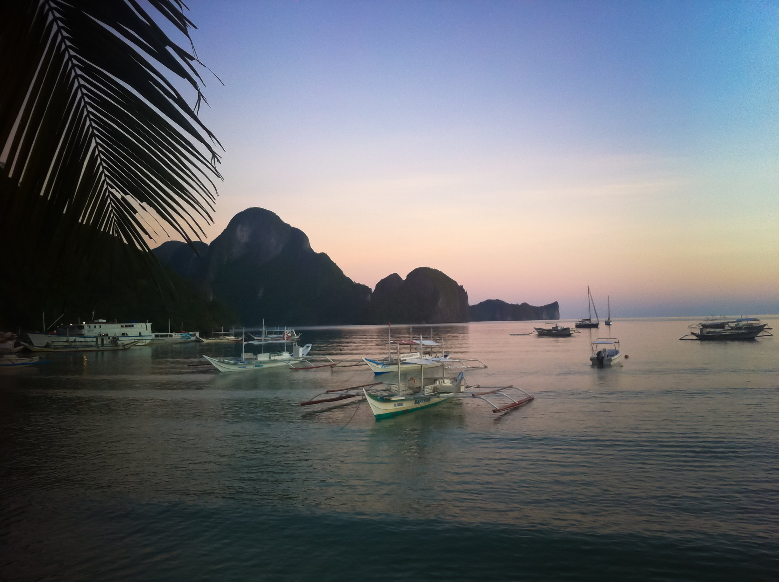 Boats anchored in calm water near a mountainous island at sunset, with a palm leaf partially visible on the left.