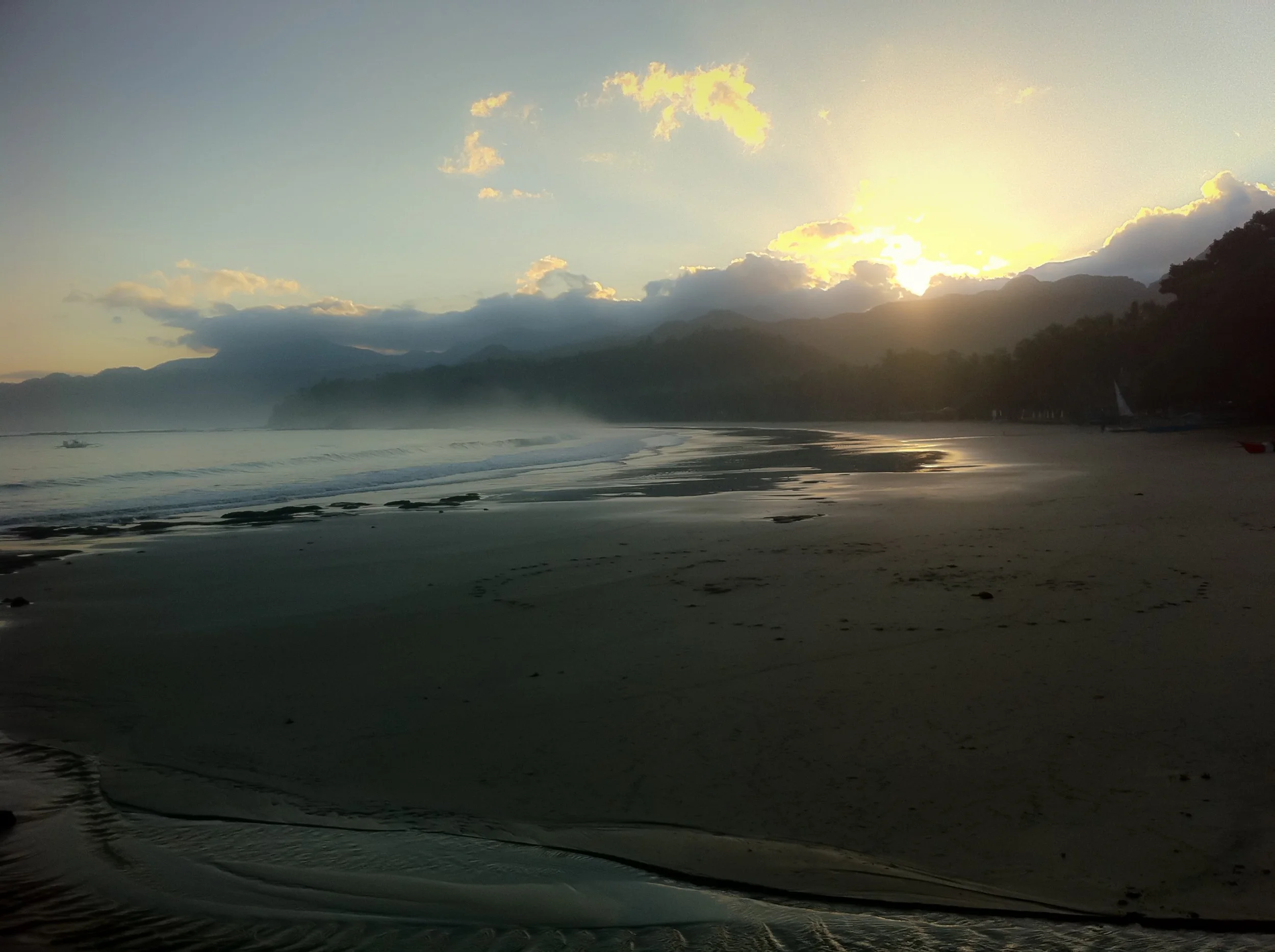 Sunset over a quiet beach with mountains in the distance, mist rising from the water, and a sailboat on the shore.
