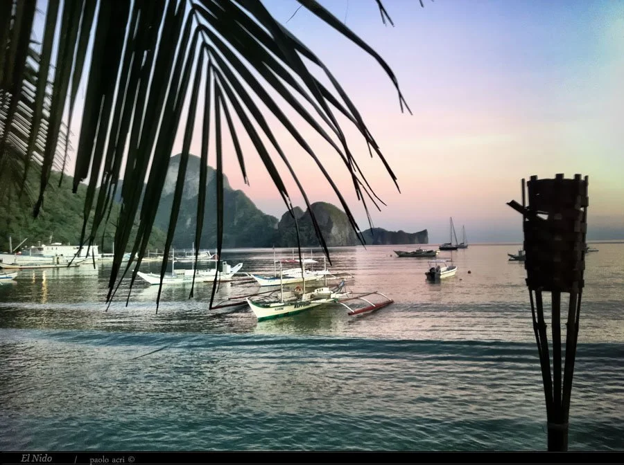 Boats anchored near the shore at sunset with mountains in the distance and a tiki torch in the foreground.
