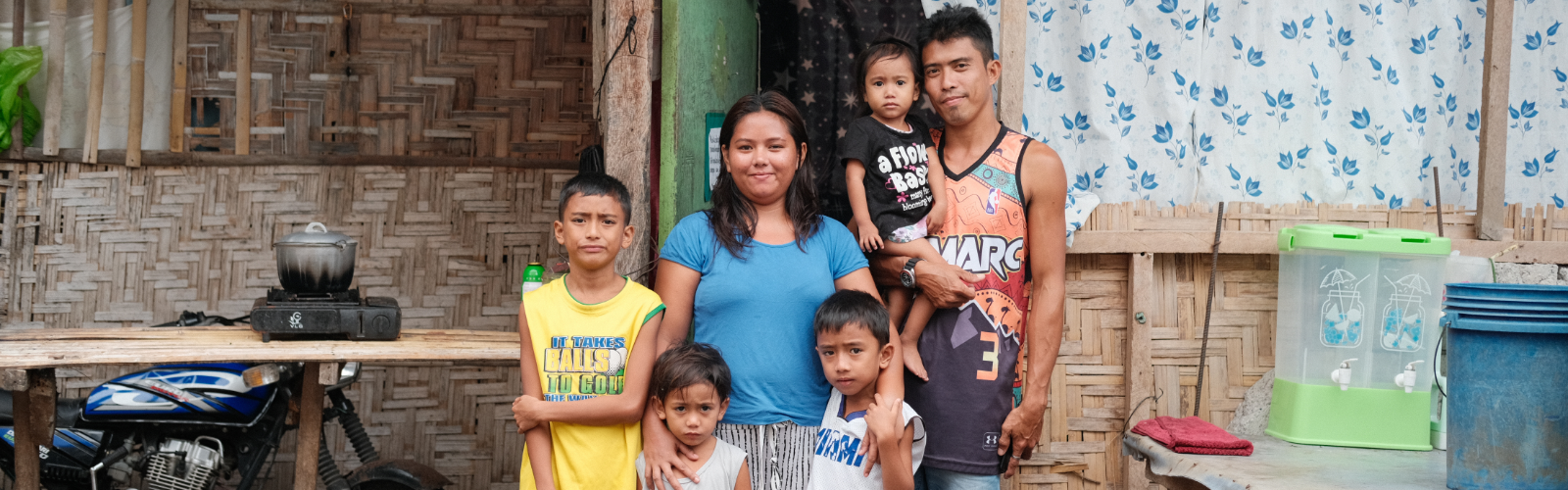 A family of seven posing outdoors in front of a woven wall, including two adults and five children, with various household items around.