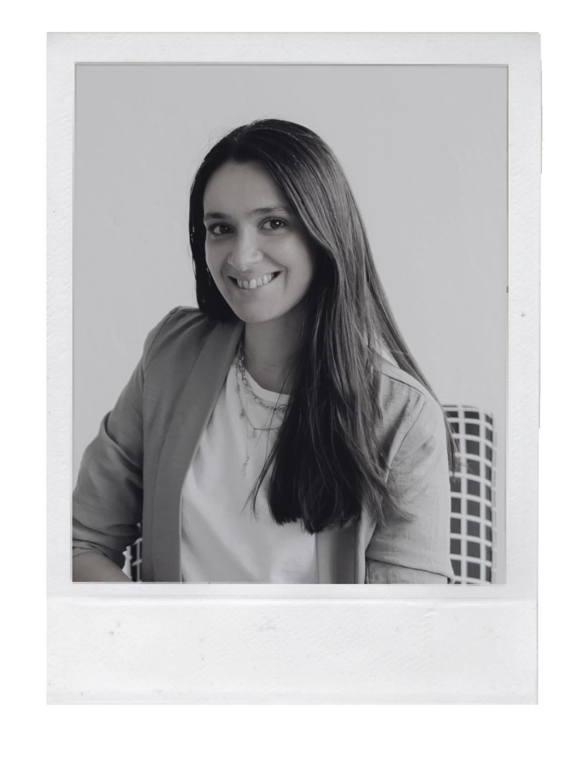 Retrato en blanco y negro de una mujer sonriendo, con cabello largo y oscuro, usando un blazer y varias cadenas, sentada en una silla con respaldo de cuadro.