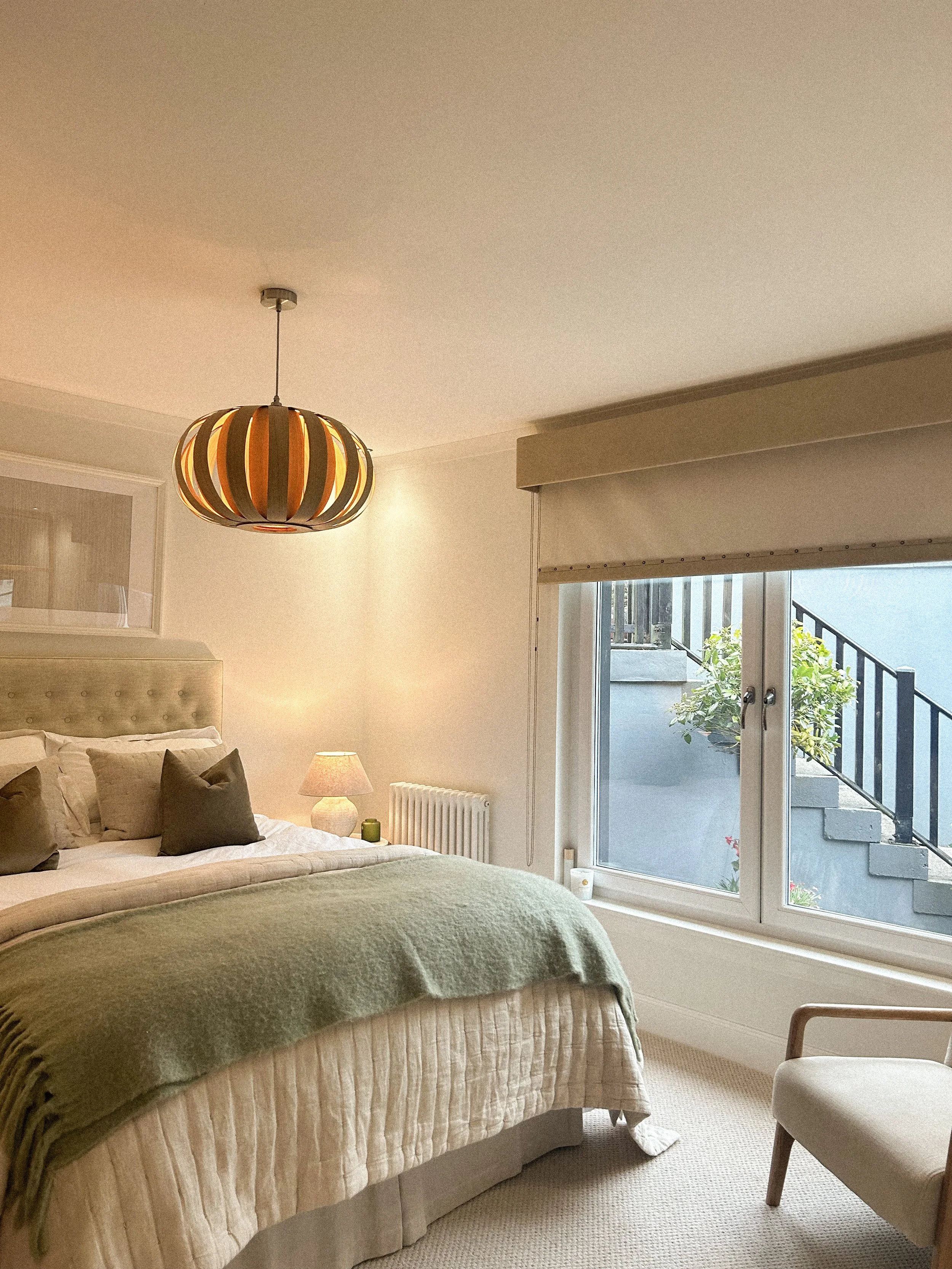 A cozy bedroom with a bed, green and beige bedding, a nightstand with a lamp, and a large window with a view of stairs outside.“Residential interior design project in Scotland by [Studio Name], working across Glasgow, Edinburgh, Dundee and the Highla