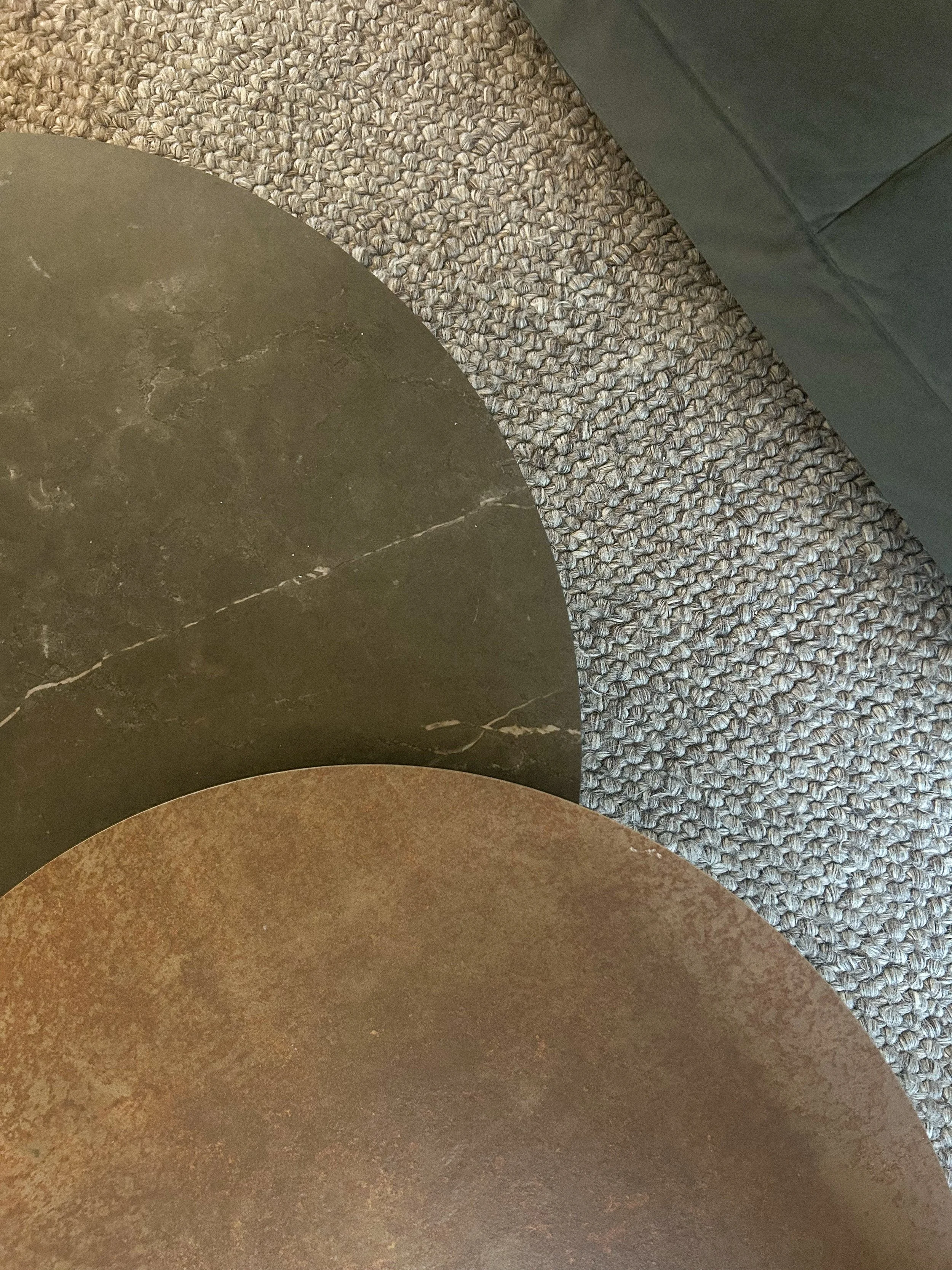 Top-down view of three overlapping round tabletops with different textures and colors: dark green with white veining, brown with a mottled pattern, and a gray textured surface, next to a gray armrest on a gray carpet.Residential interior design proje