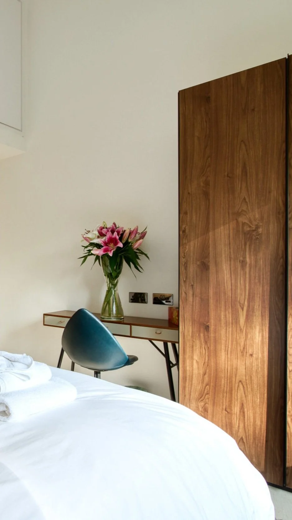 A bedroom with a white bed, a bedside table with pink lilies in a glass vase, a wooden wardrobe, and a black chair.Residential interior design project in Scotland - Interior Designer Scotland - Interiors 
