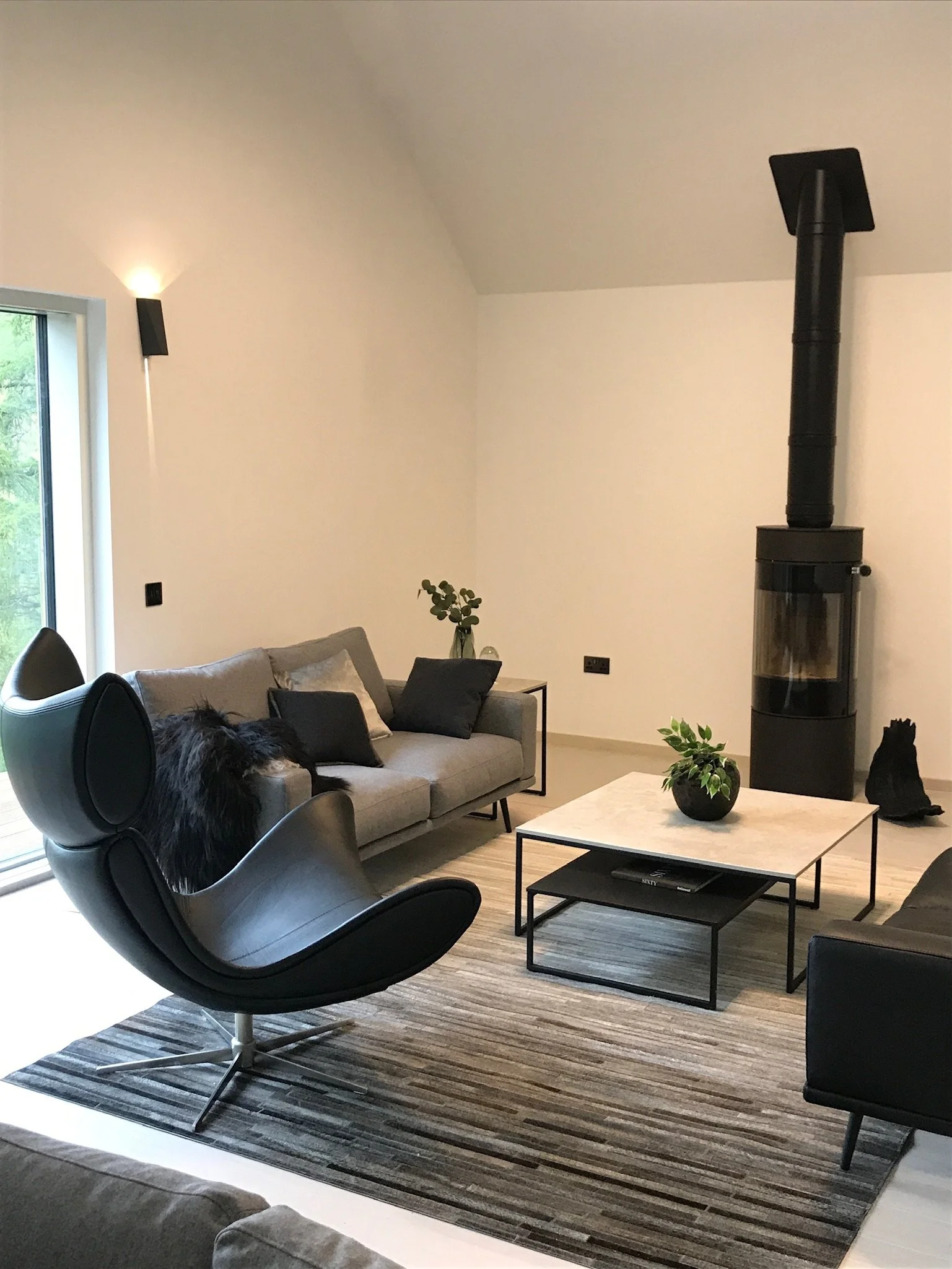 Modern living room with a light gray sofa, black and gray pillows, a black lounge chair, a white coffee table with a green plant, a black wood stove, a side table with a vase of toiletries, and a large window with greenery outside.Residential interio