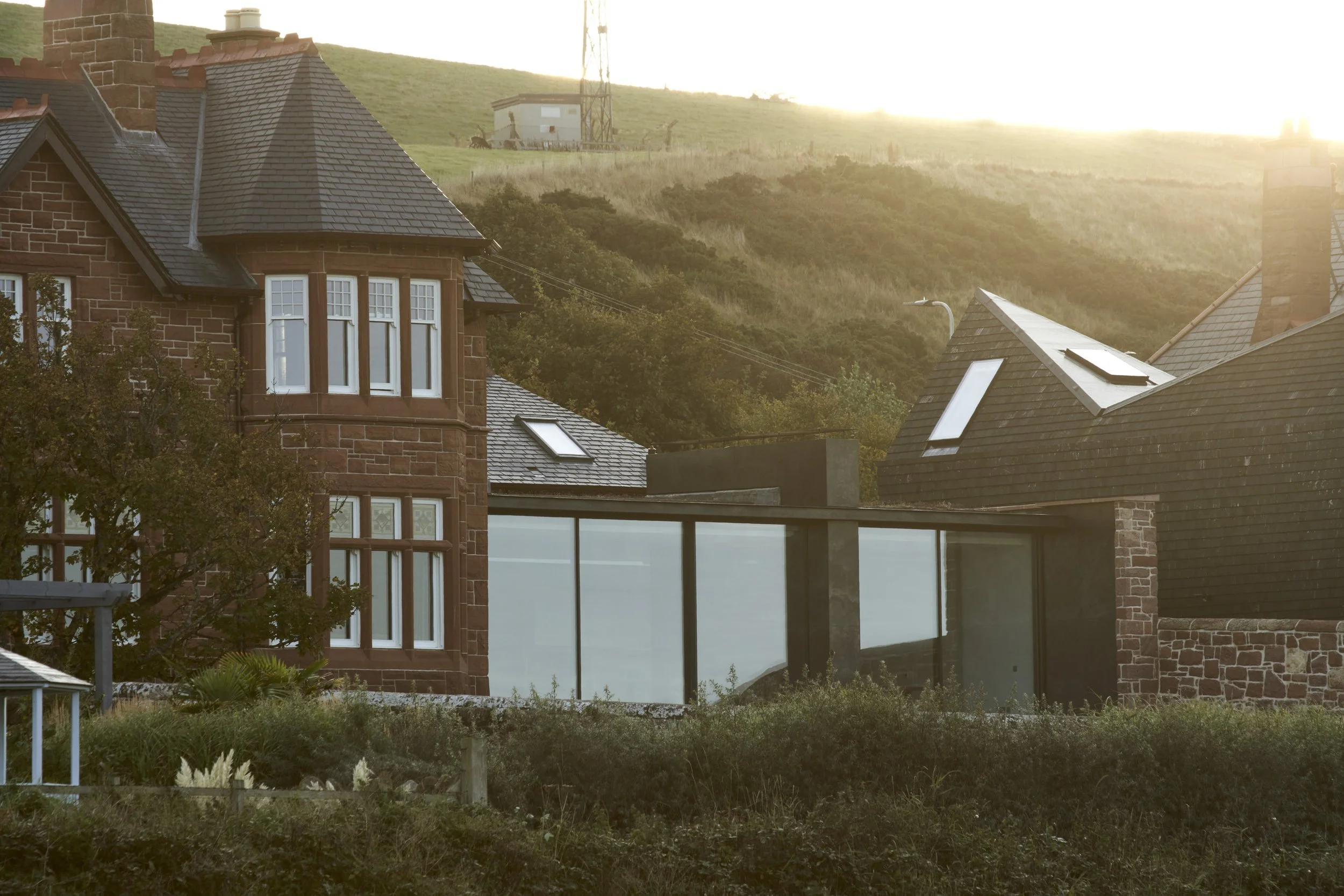 Modern house with large glass windows, brick and stone exterior, surrounded by greenery, with hills in the background and the sun setting.Residential interior design project in Scotland - Interior Designer Scotland - Interiors 