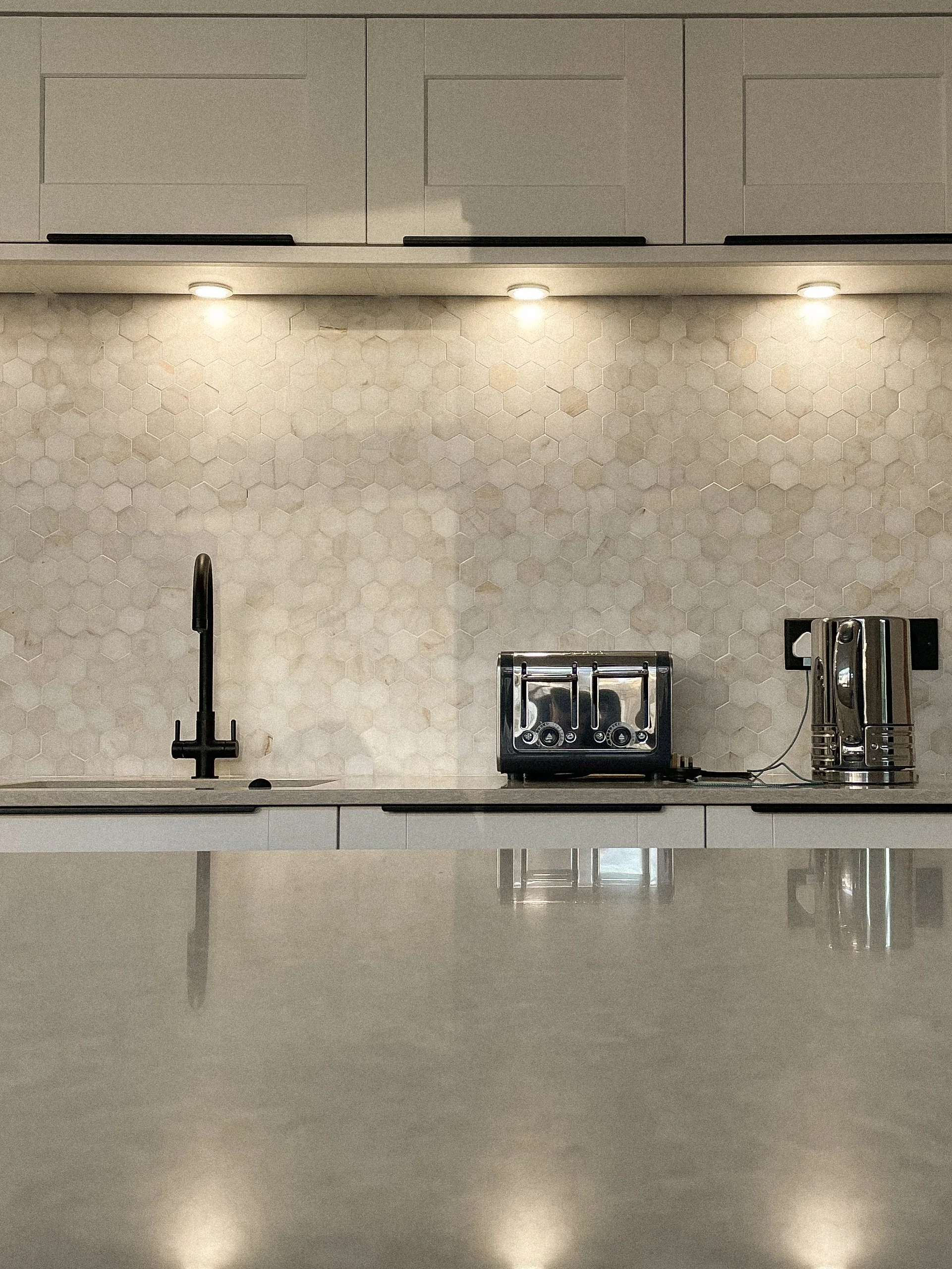 Minimalist kitchen with white cabinetry, hexagonal tiled backsplash, black faucet, black toaster, and silver electric kettle under ceiling-mounted lights.“Residential interior design project in Scotland by [Studio Name], working across Glasgow, Edinb