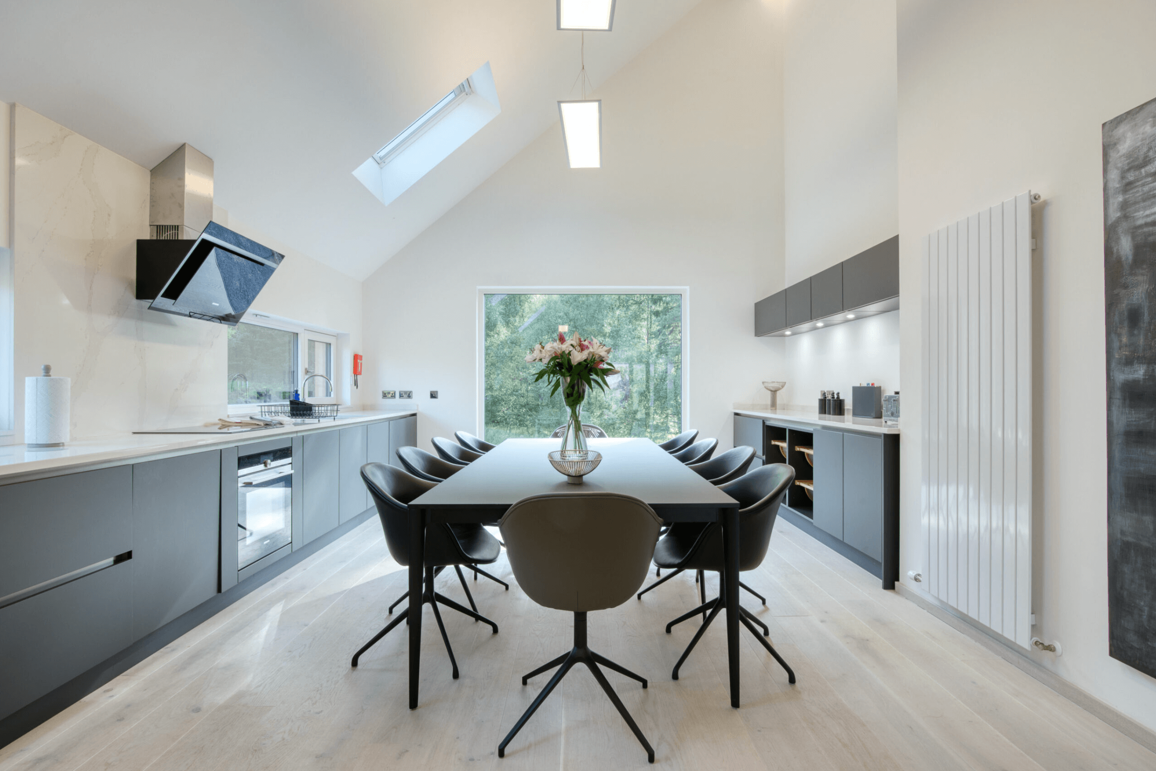 Modern kitchen with a long dining table, black chairs, a vase of flowers, large window showing trees, sleek cabinets, and skylights.Residential interior design project in Scotland - Interior Designer Scotland - Interiors 