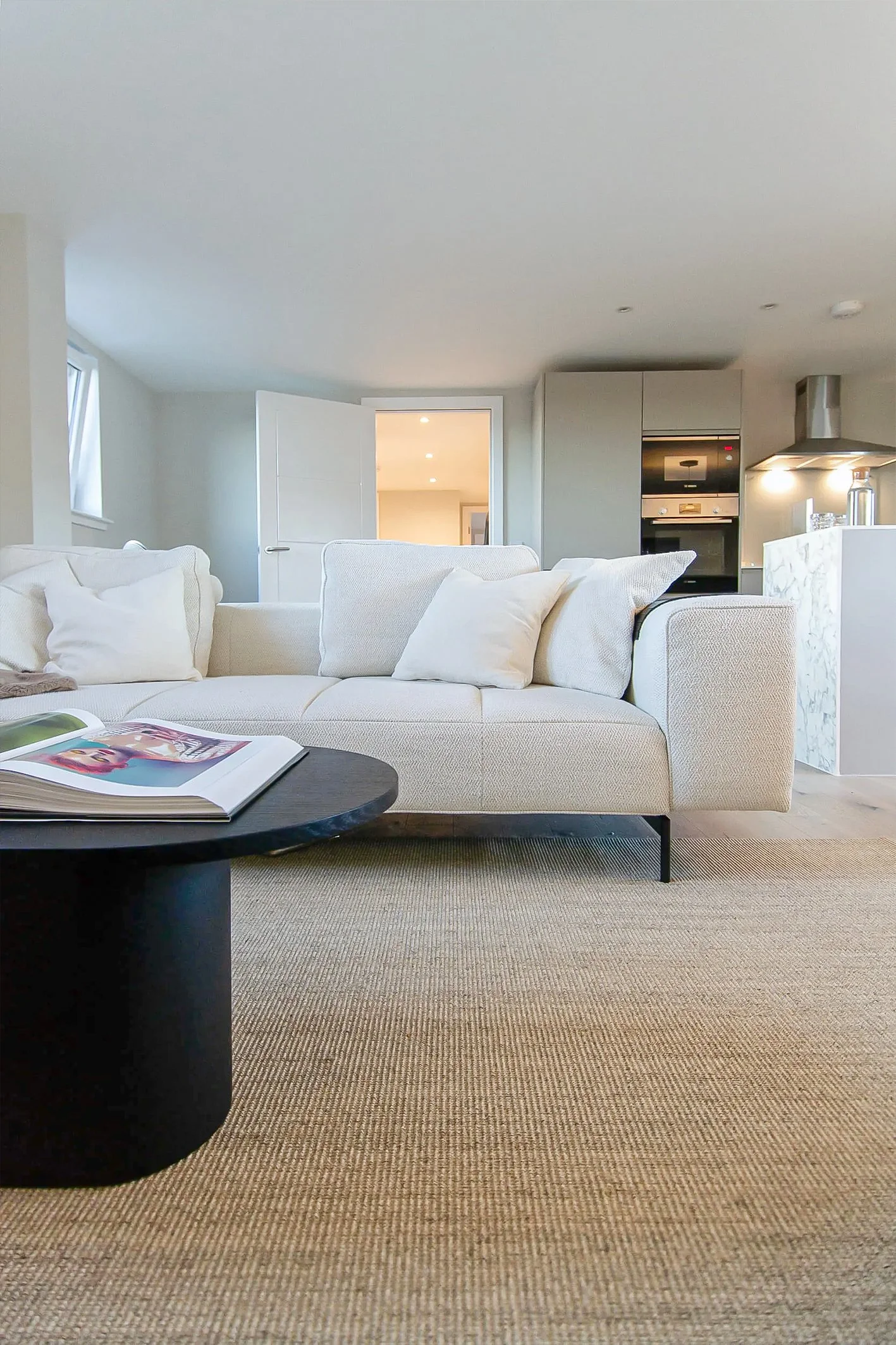 A modern living room with a beige sofa, white pillows, a round black coffee table with magazines, a textured beige carpet, and a contemporary open kitchen with a marble island and built-in oven in the background.Residential interior design project in