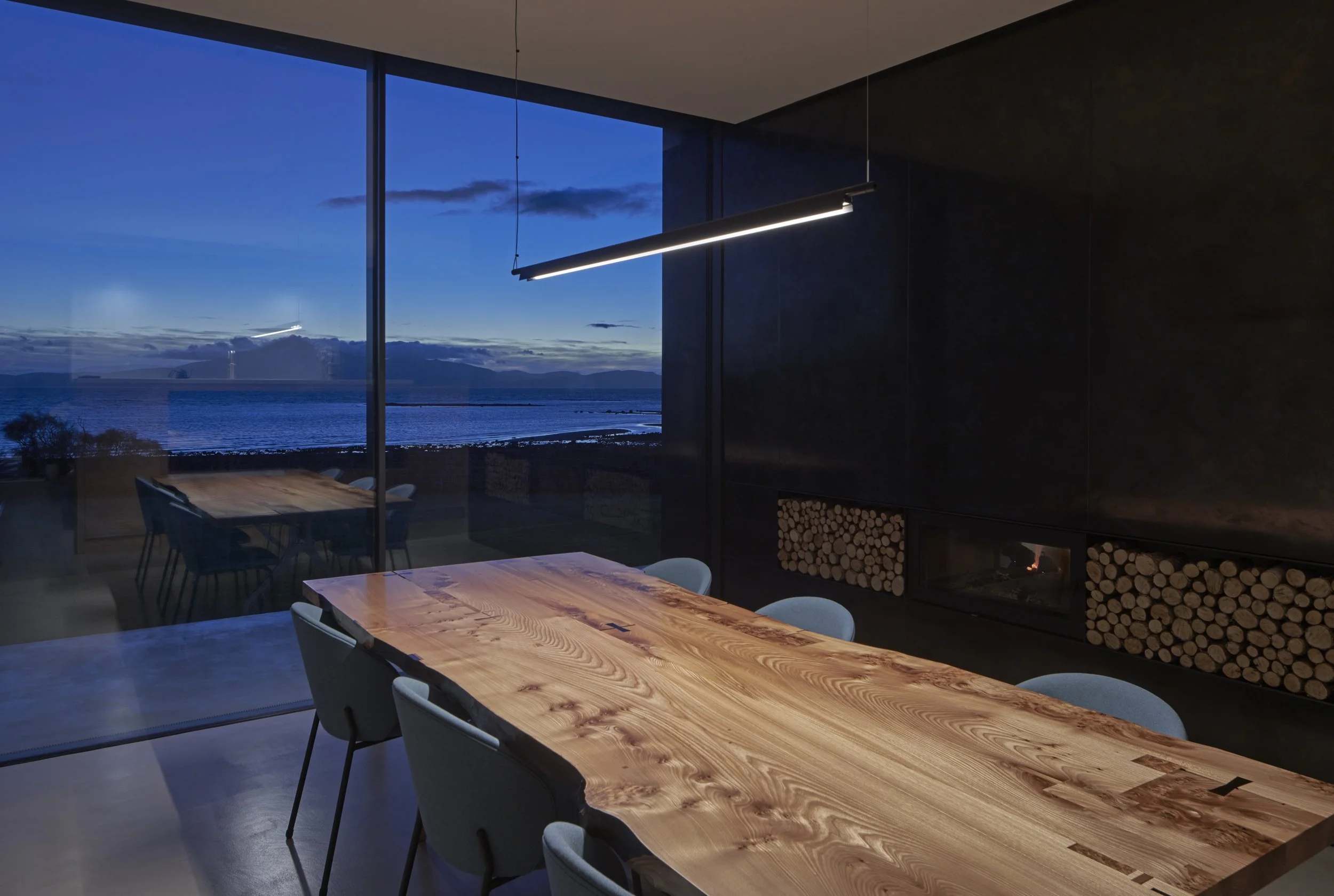 Modern dining room with a large wooden table, surrounded by gray chairs, overlooking a waterfront view through large glass windows at dusk. There is a sleek pendant light above the table and a fireplace with stacked logs on the dark wall.Residential 