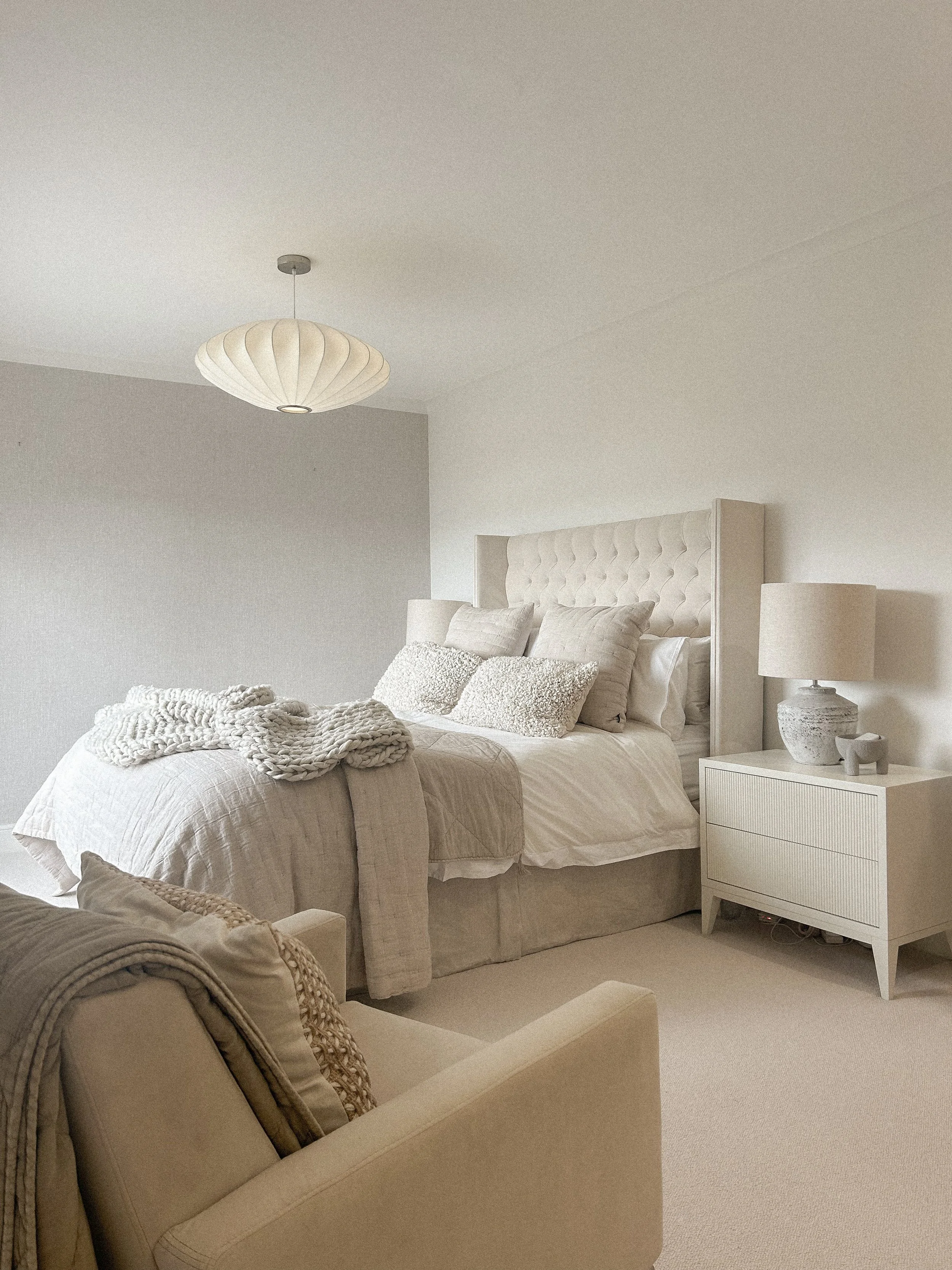 A cozy bedroom with a white upholstered bed, multiple pillows, a beige armchair, and a nightstand with a lamp, all in a neutral color palette.“Residential interior design project in Scotland by [Studio Name], working across Glasgow, Edinburgh, Dundee