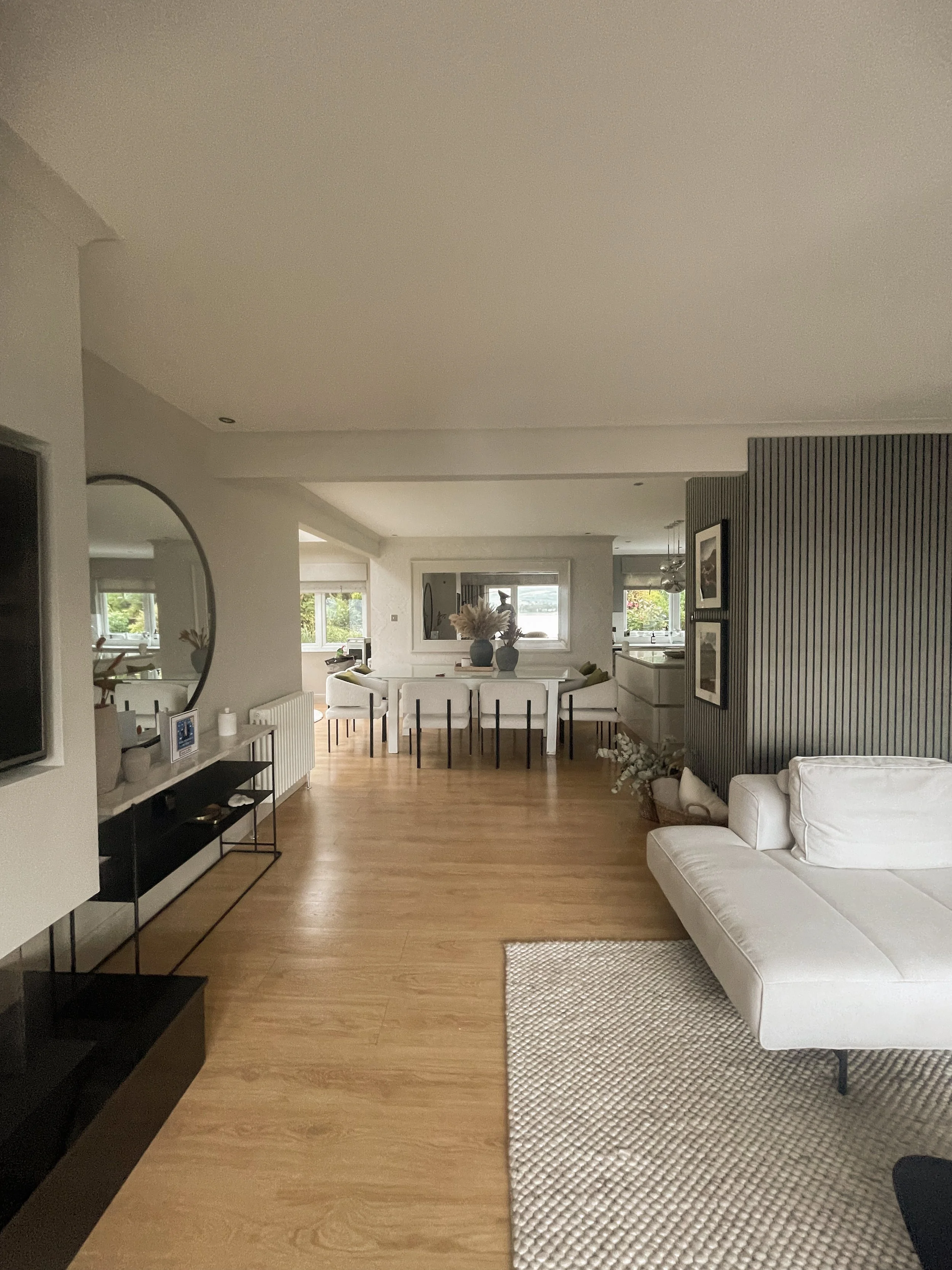 A spacious, modern living room with light wood flooring, a white sofa, a black TV stand, a round mirror, and a dining area with white chairs and a table decorated with vases and flowers.“Residential interior design project in Scotland by [Studio Name