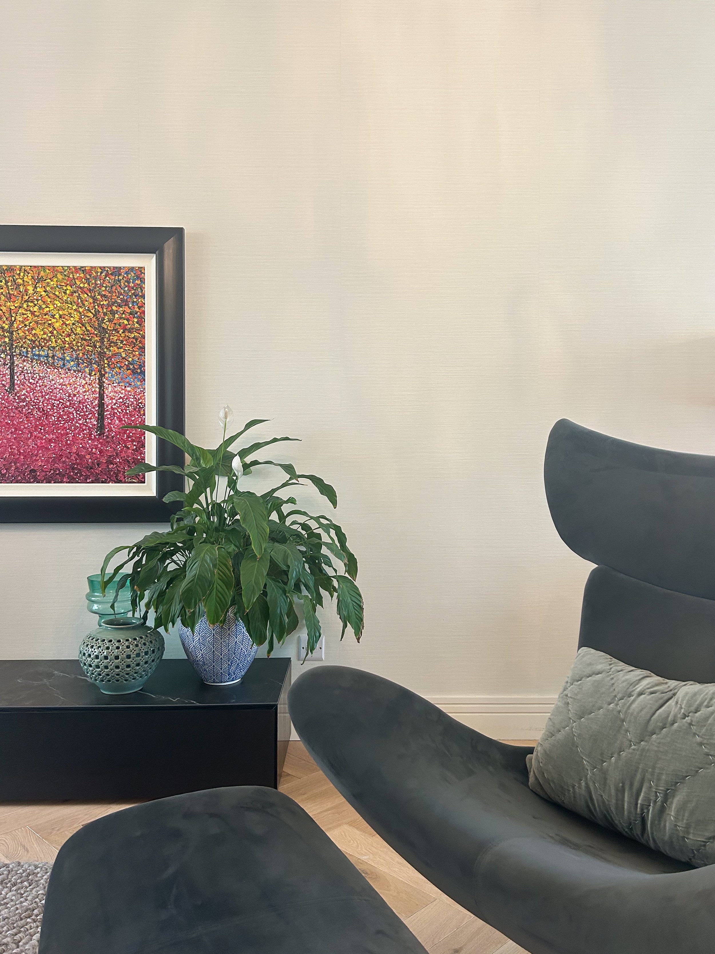 A living room corner with a black leather chair, a dark side table holding a potted plant, and decorative pottery, with a framed colorful abstract painting on a cream-colored wall.Residential interior design project in Scotland - Interior Designer Sc