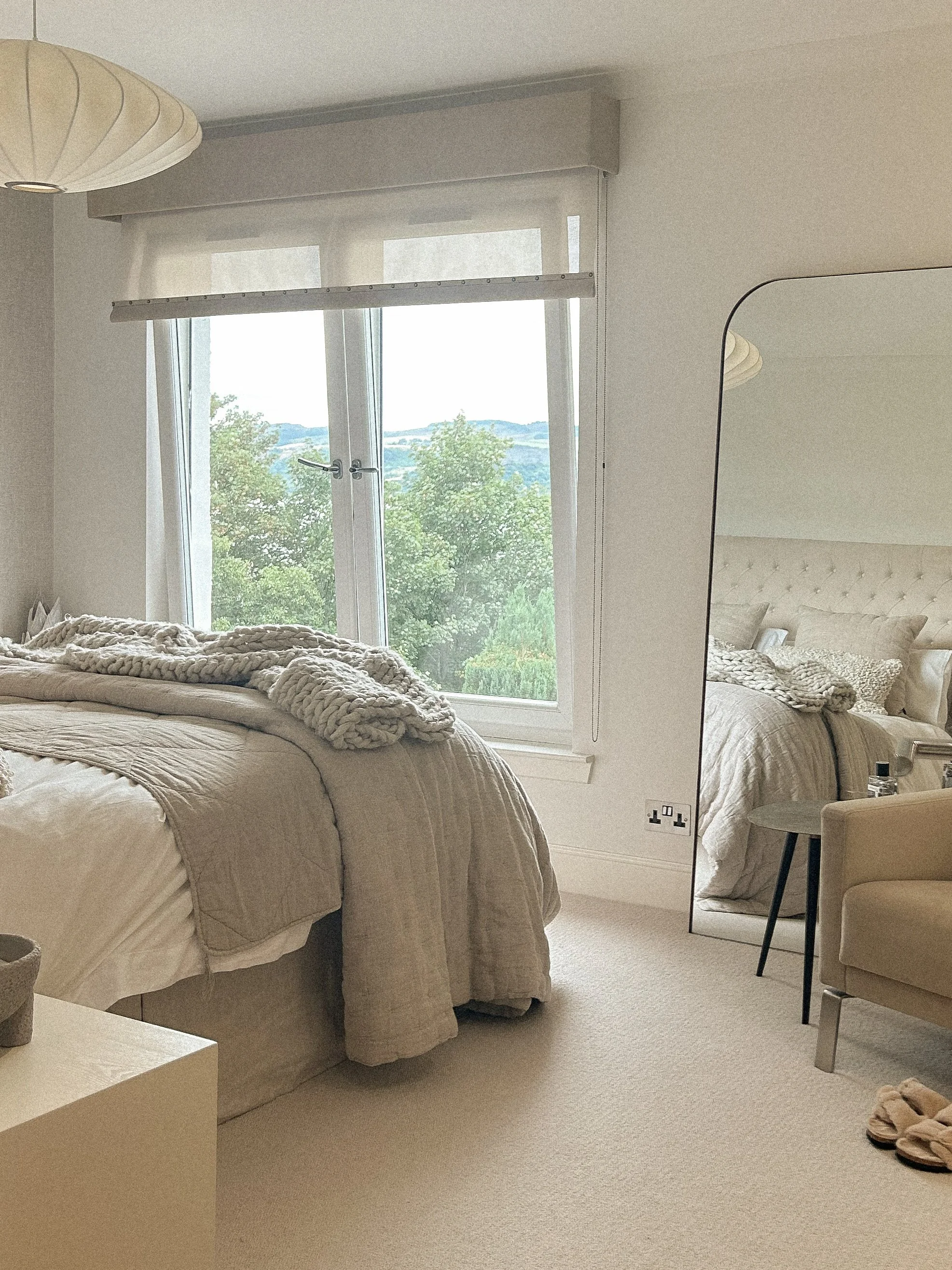 A bedroom with a bed covered in cream-colored bedding, a large window showing trees and hills outside, and a full-length mirror reflecting part of the room.“Residential interior design project in Scotland by [Studio Name], working across Glasgow, Edi
