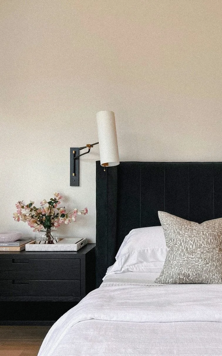 A bedroom with a black headboard, white bedding, and a decorative pillow. Beside the bed, a black nightstand holds a vase with pink flowers and a tray with books. A white wall-mounted light fixture is above the headboard.Residential interior design p