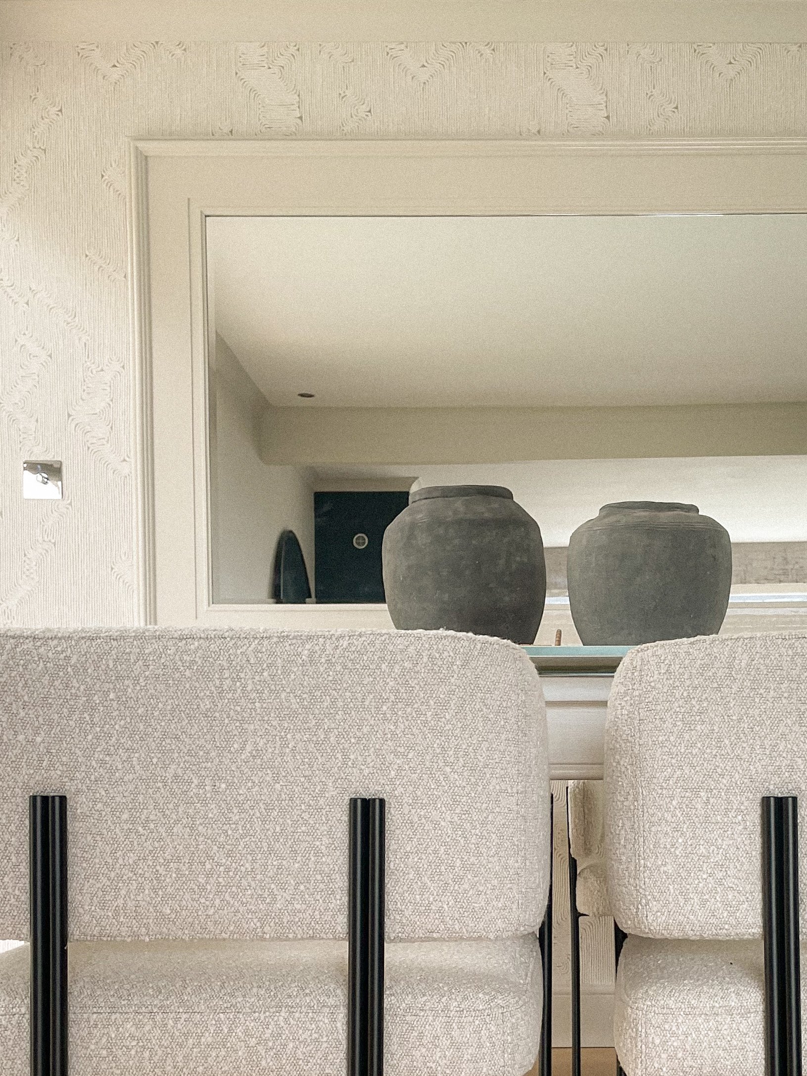Close-up of two beige upholstered chairs with black metal legs, facing a mirror that reflects two dark ceramic vases on a white surface, in a neutral-colored room.“Residential interior design project in Scotland by [Studio Name], working across Glasg