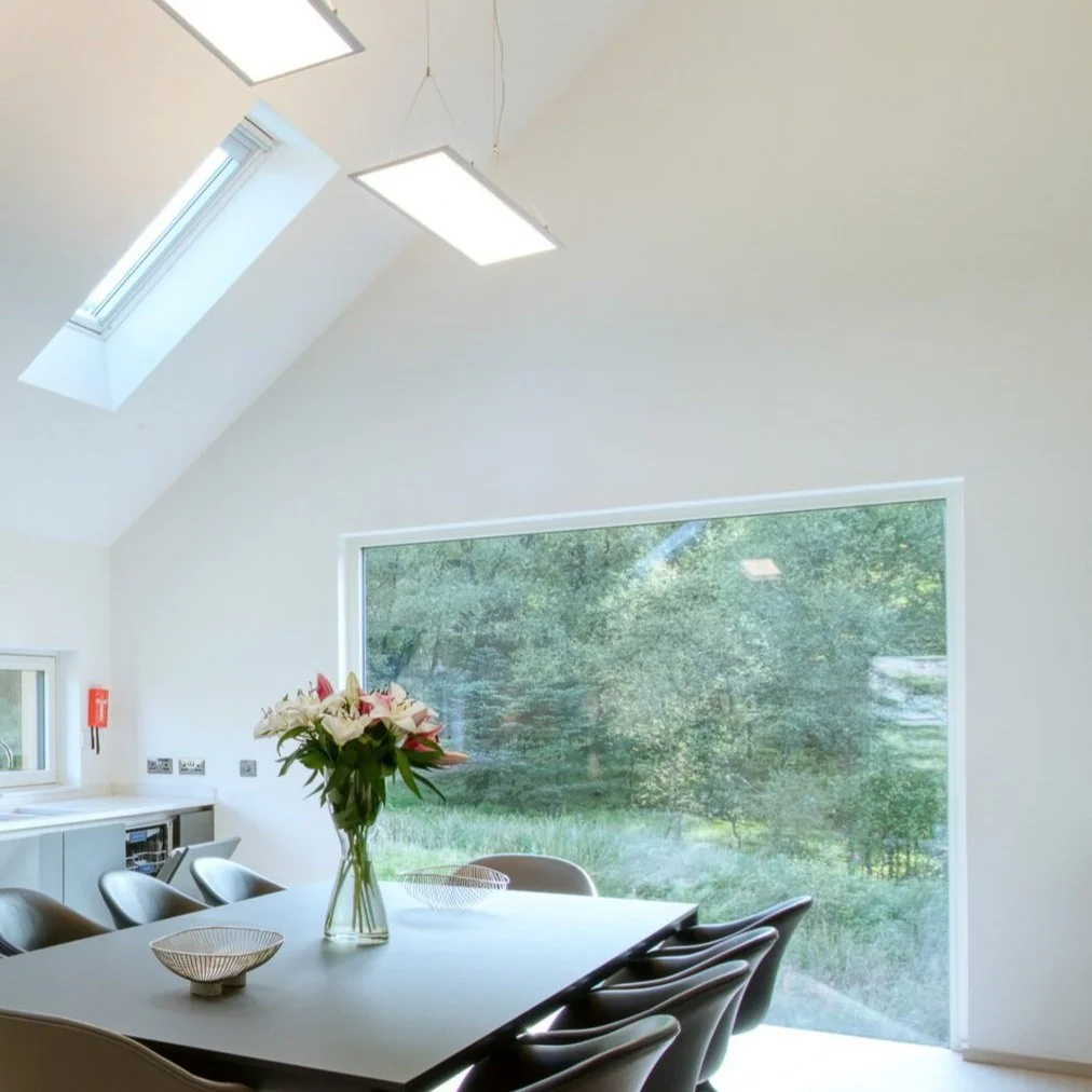 Modern dining room with a large window overlooking greenery, a white table with a flower vase, and black chairs, illuminated by ceiling lights and a skylight.Residential interior design project in Scotland - Interior Designer Scotland - Interiors 