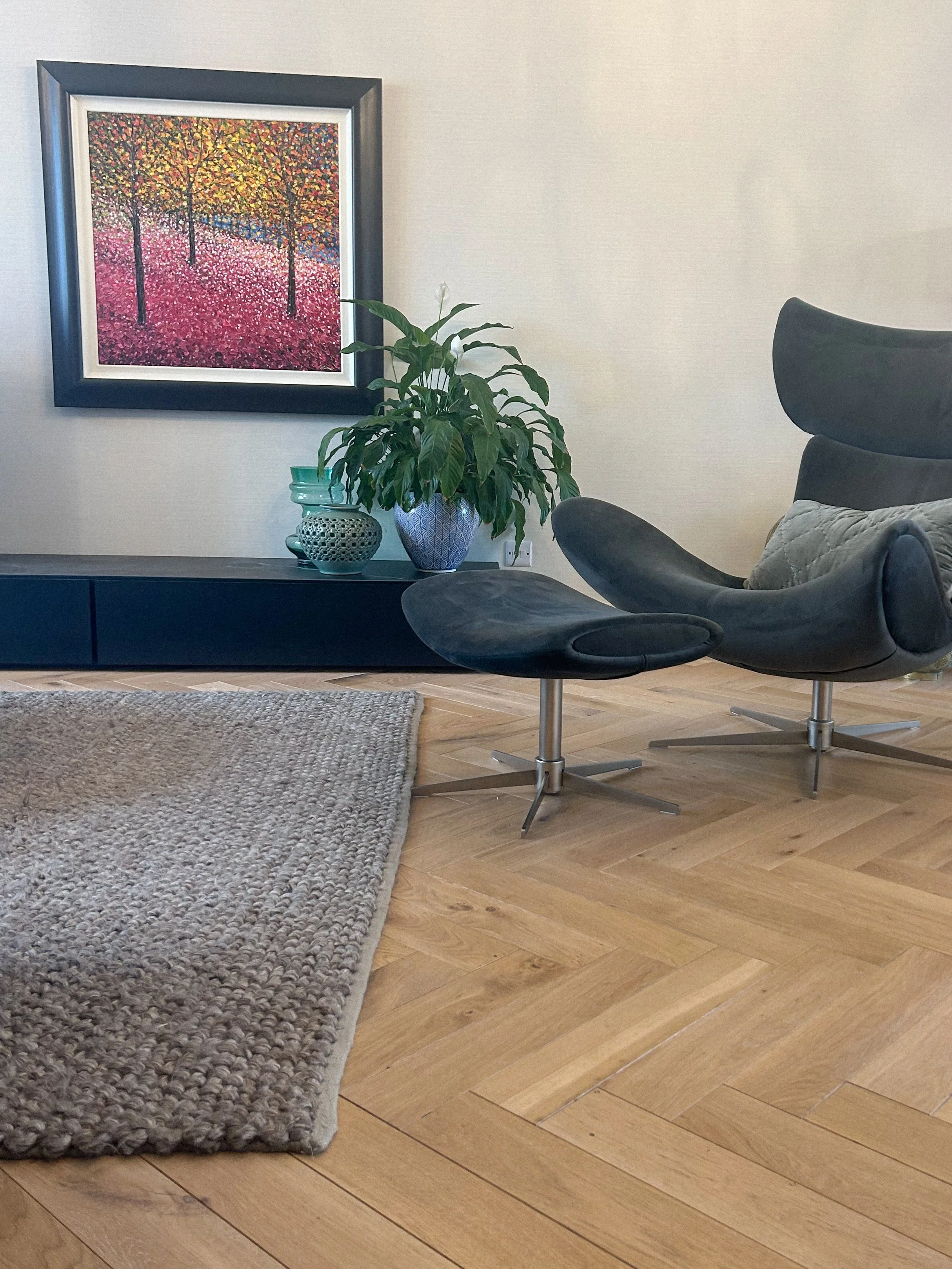 Modern living room corner with a black leather lounge chair and matching ottoman, a black low cabinet with decorative vases, a green houseplant, a colorful painting of trees with fall foliage on the wall, hardwood floor and a textured beige rug.Resid