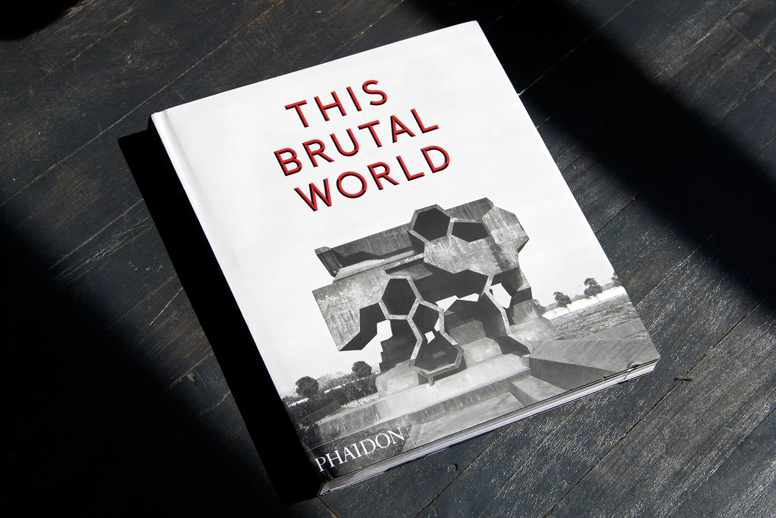 A book titled 'This Brutal World' by Phaidon is placed on a dark wooden surface. The cover features a black and white photo of a Brutalist building with geometric concrete shapes.Residential interior design project in Scotland - Interior Designer Sco