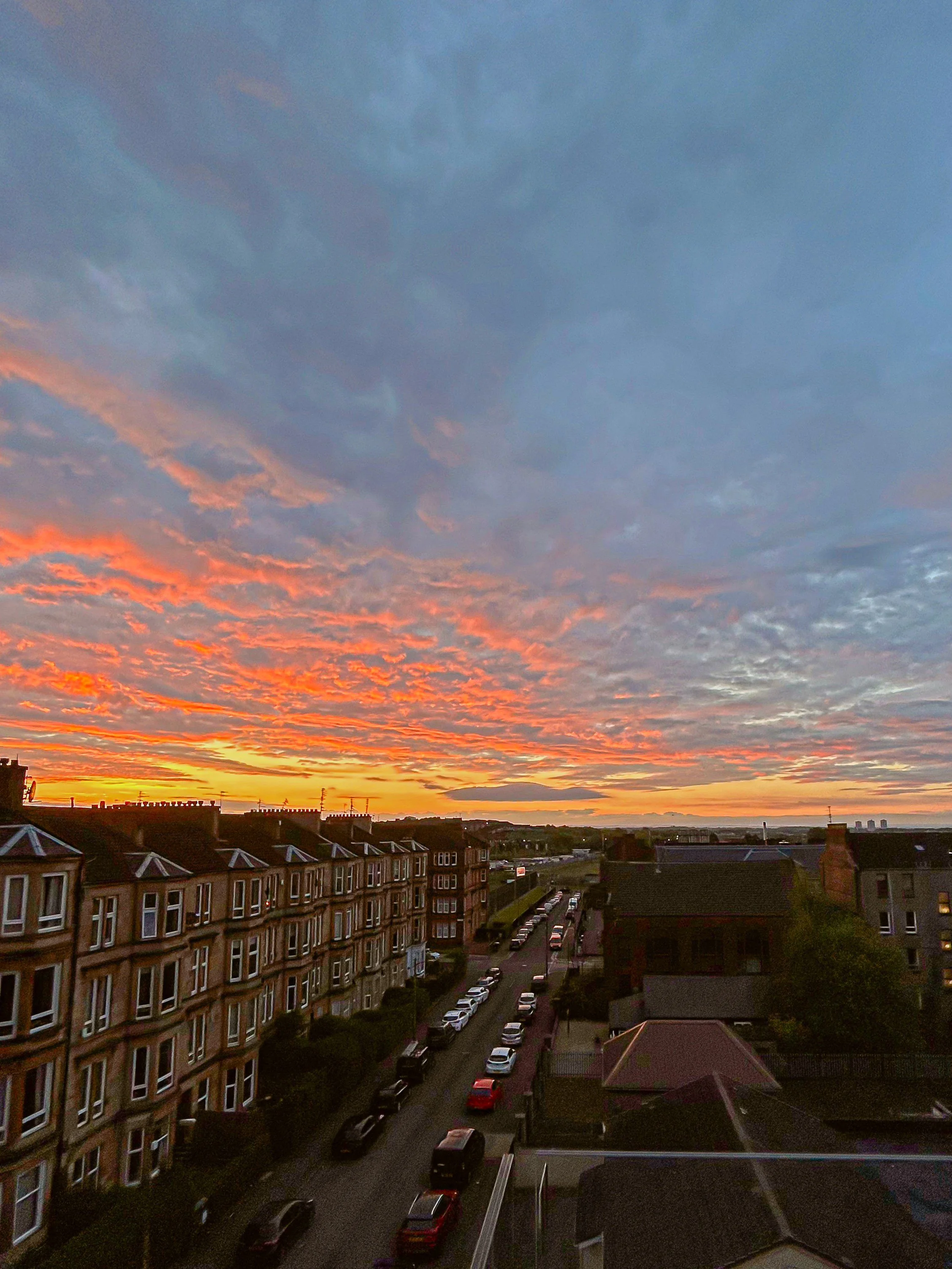 A sunset over a residential area with colorful orange and purple clouds in the sky, apartment buildings, and a line of parked cars along the street.Residential interior design project in Scotland - Interior Designer Scotland - Interiors 